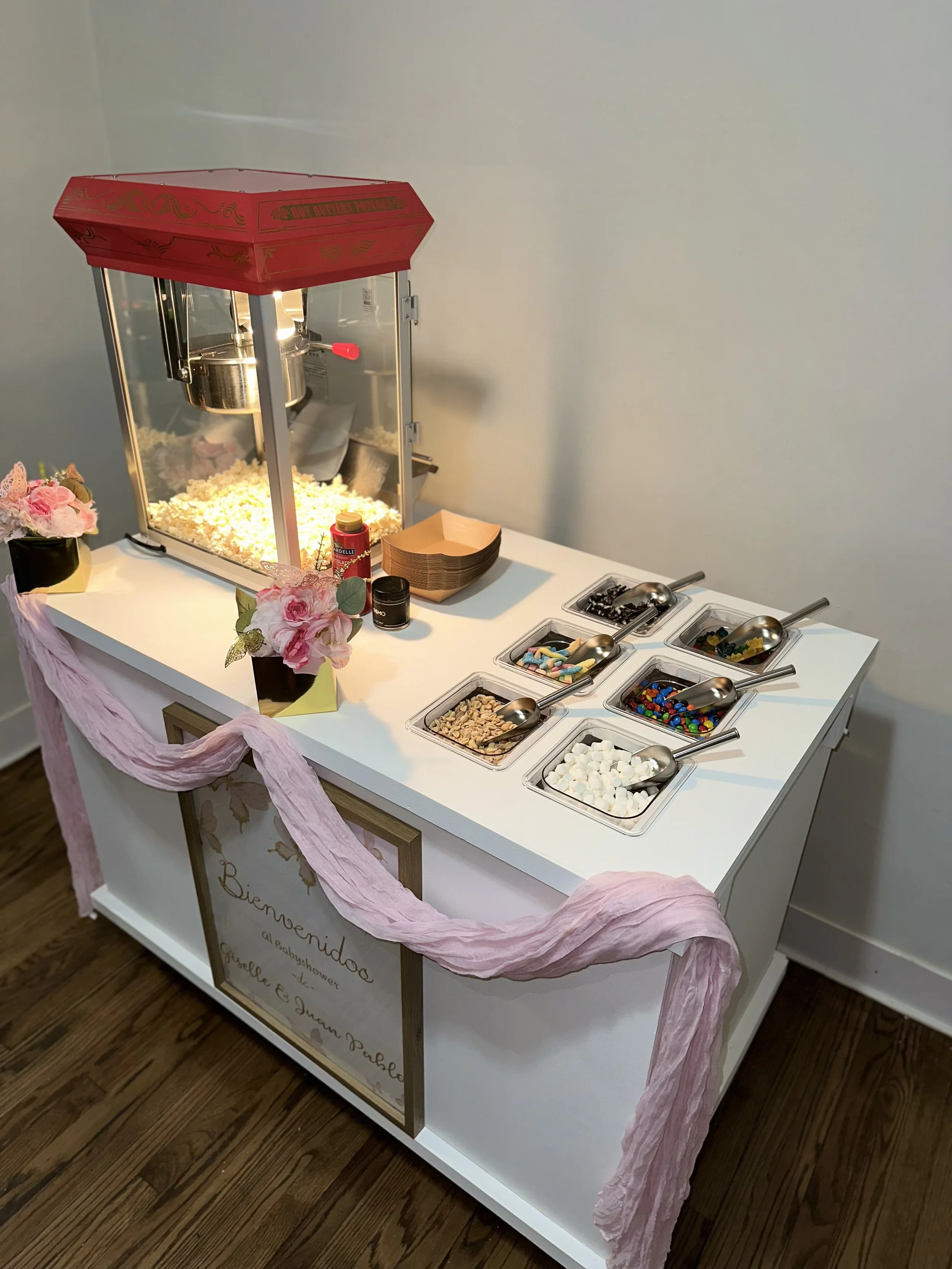 A popcorn machine with a glass enclosure on a white table, decorated with pink flowers and a pink fabric drape. The table has small containers of toppings and candies, and a wooden sign with welcoming message, on a brown wooden floor.