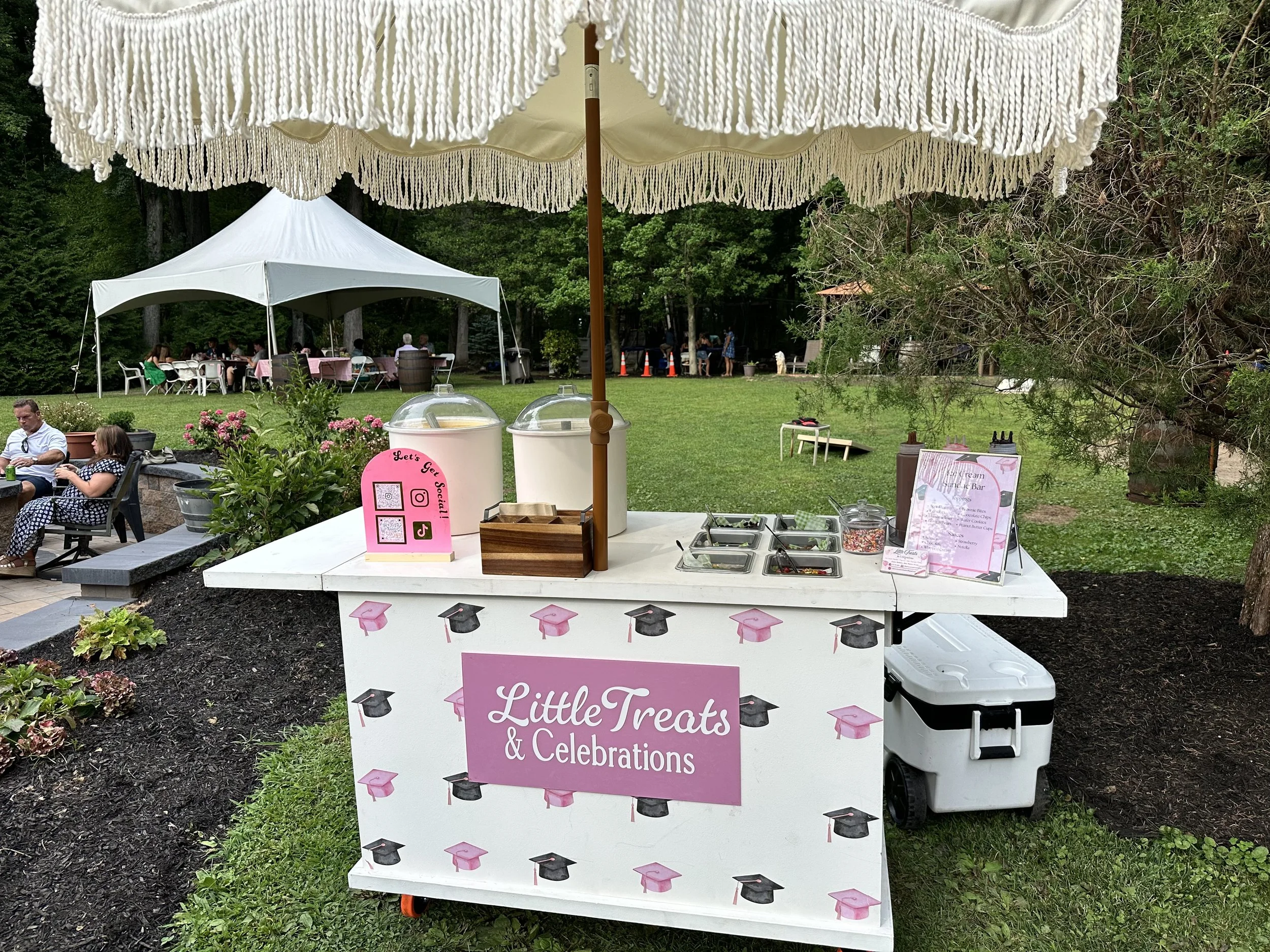 A white dessert and treat table with pink and black graduation cap decorations, set outside with two large umbrellas, multiple food containers, and a pink sign that says "Little Treats & Celebrations." In the background, there are tents and people ga