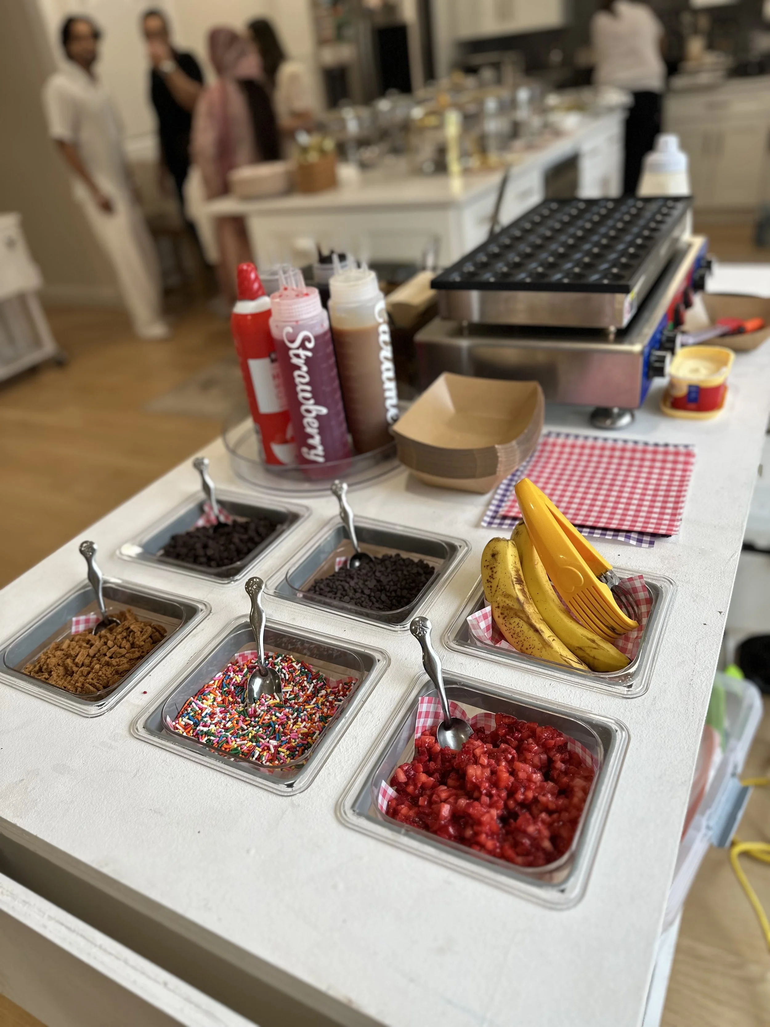 A dessert or snack bar with toppings including sprinkles, chocolate chips, and fruit, set up on a white countertop with people in the background at a social gathering.