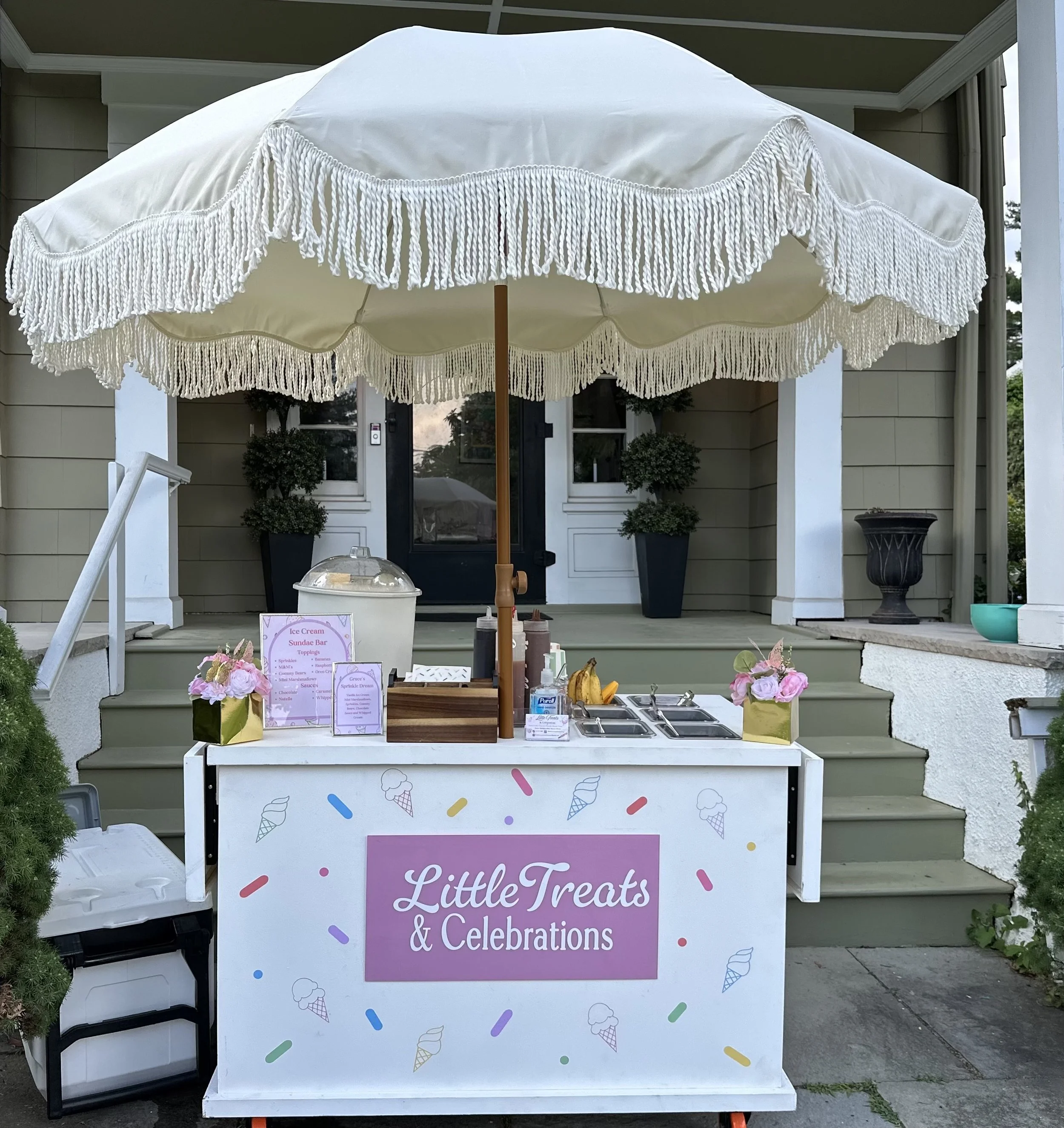 Ice cream stand with colorful sprinkles pattern and a pink sign that reads "Little Treats & Celebrations". The stand has a large white umbrella with tassels, a sign listing ice cream flavors, tubs of toppings, flowers, and a container for napkins and
