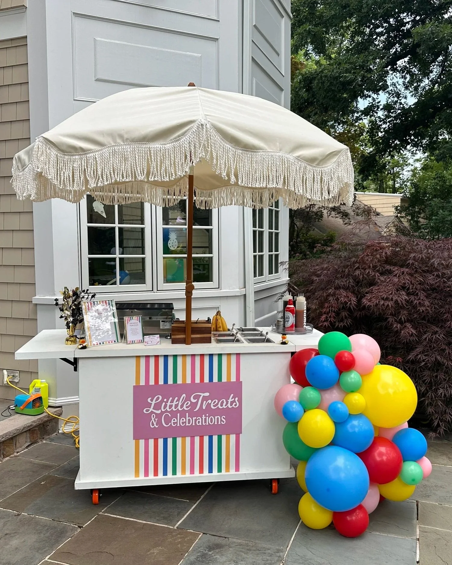 A decorated outdoor party stand with a sign that reads 'Little Treats & Celebrations', colorful balloons on the right side, and a white umbrella casting shade over the setup. The stand includes various party supplies and is set against a house with a window and trees in the background.