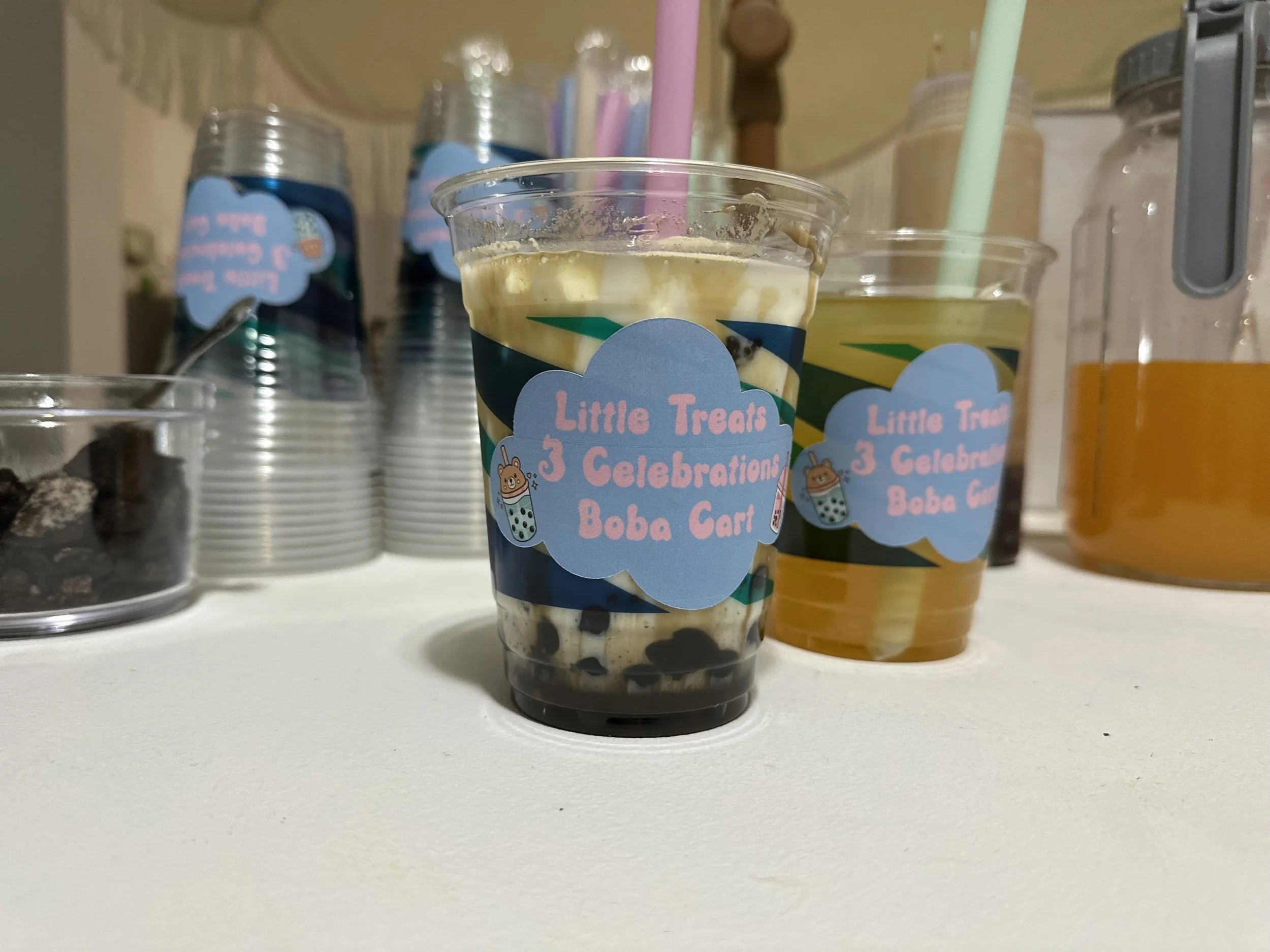 Clear plastic cup filled with boba tea topped with yellowish cream, with a pink straw. In the background, there are other cups, jars, and containers with drinks and toppings on a counter.