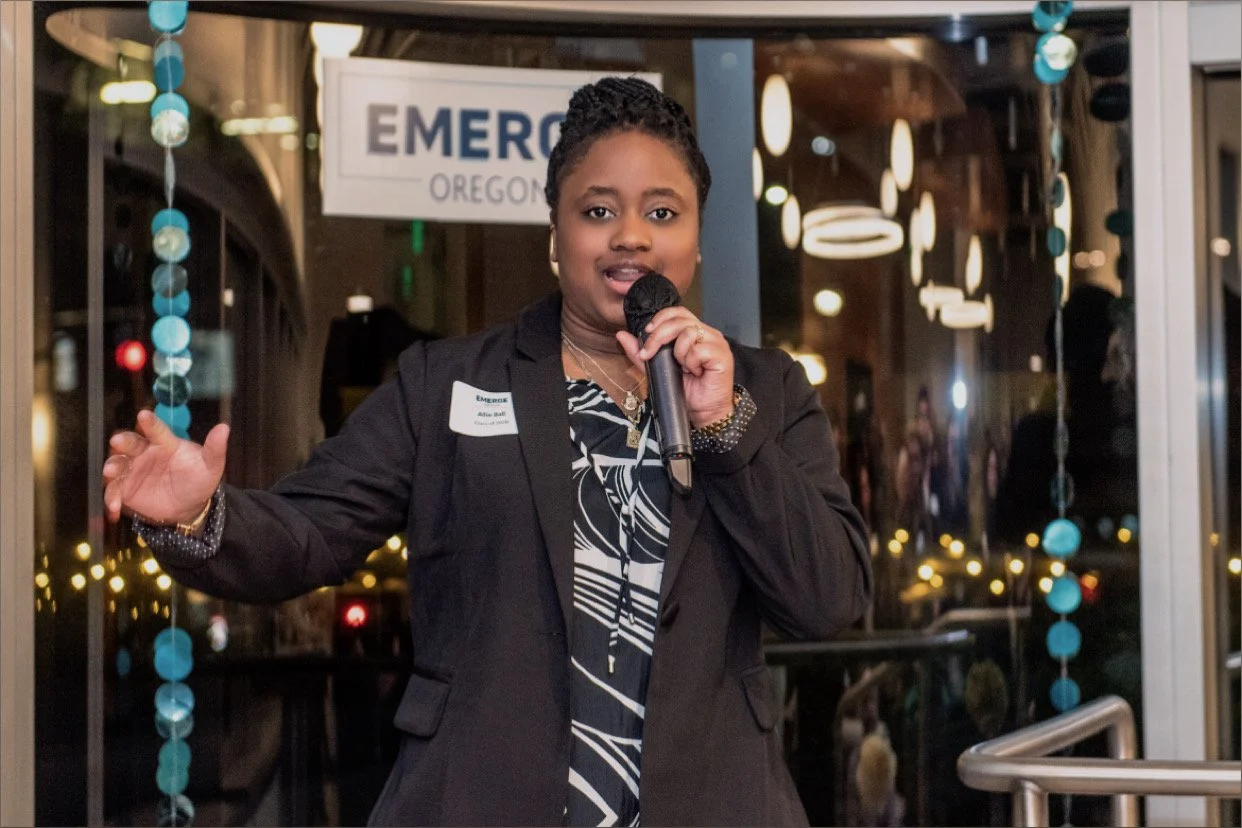 A woman speaking into a microphone at an event, standing in front of a glass door with a sign that reads 'EMERGE OREGON.' She is wearing a black blazer over a patterned top, with a name tag, and appears to be delivering a speech or presentation.