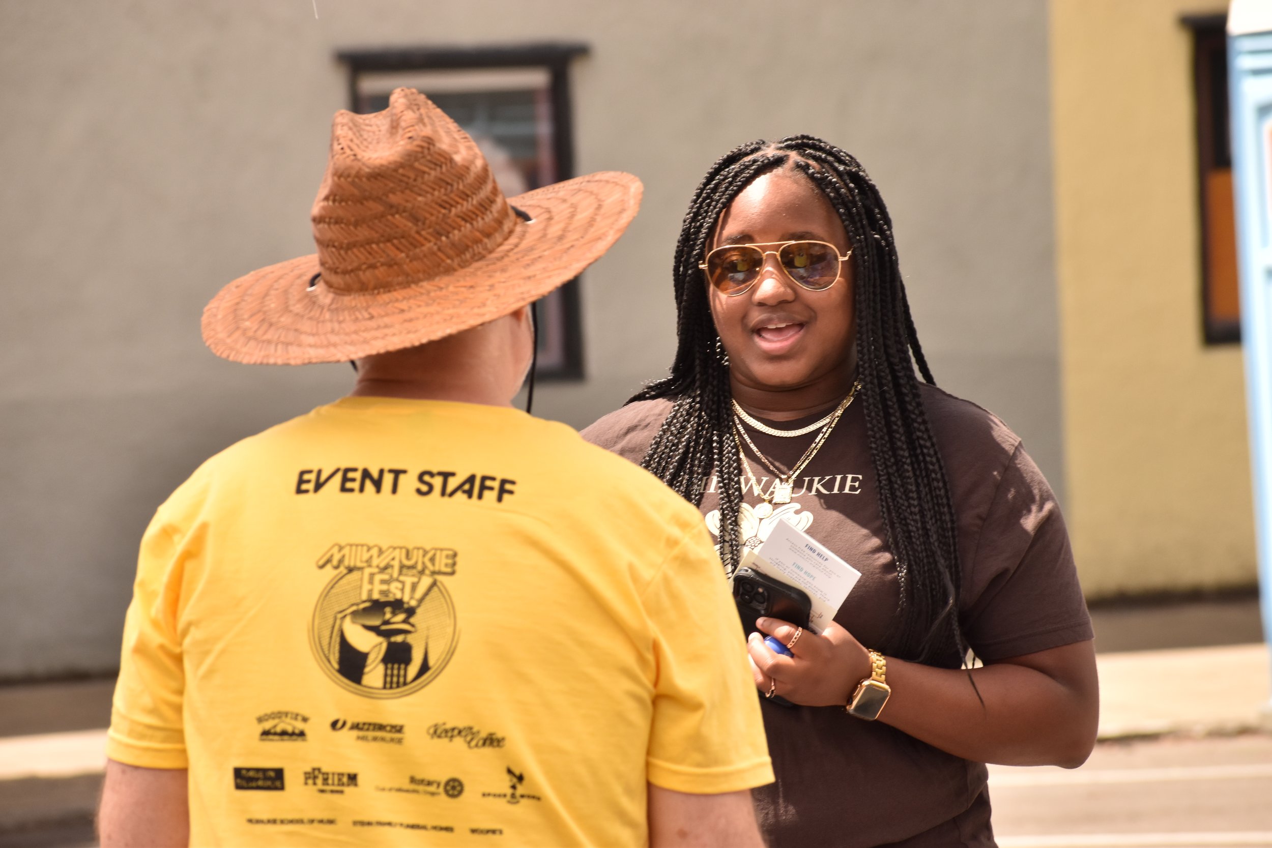 Two women engaged in conversation indoors. One woman is seen from behind, wearing a yellow shirt with "Event Staff" printed on the back and a large straw hat. The other woman, facing the camera, has long braided hair, sunglasses, and is wearing a dark T-shirt with white text. She is holding a nametag or card and has jewelry, including a necklace and bracelet.