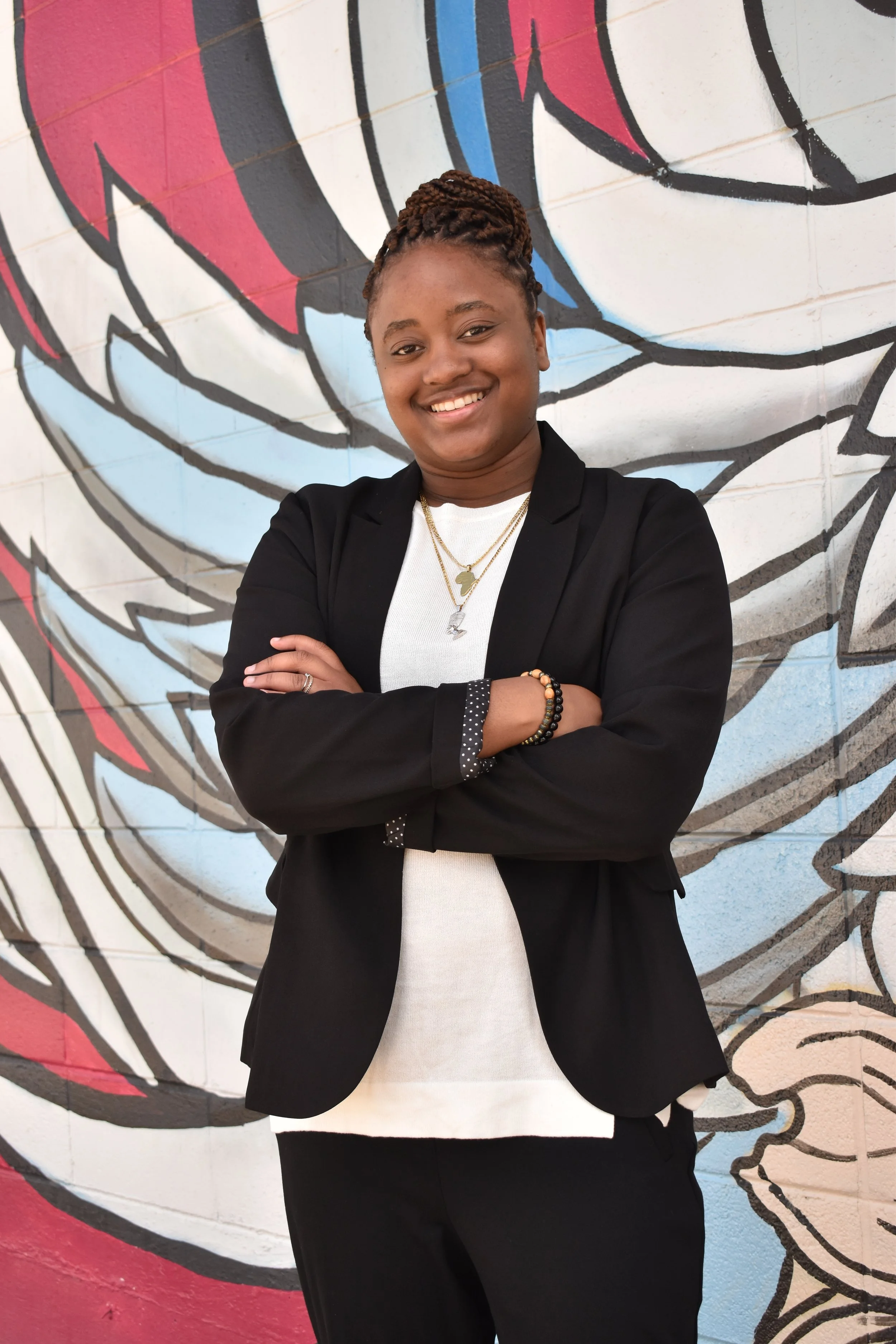 Portrait of a smiling woman with braided hair standing in front of a colorful graffiti mural on a brick wall, wearing a black blazer over a white top, with layered necklaces and bracelets.