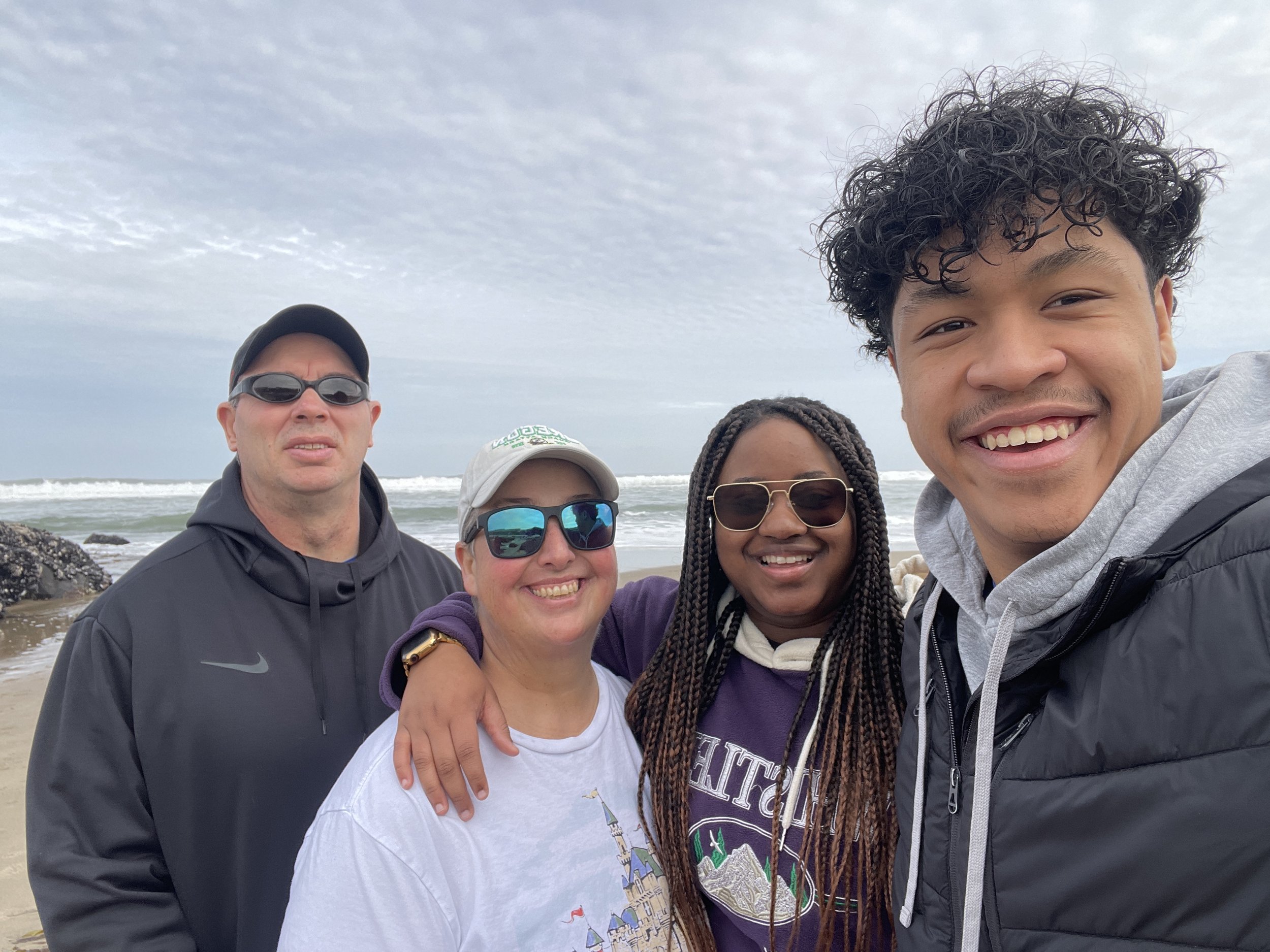 Group of five people smiling at the camera with the beach and ocean in the background, cloudy sky overhead.