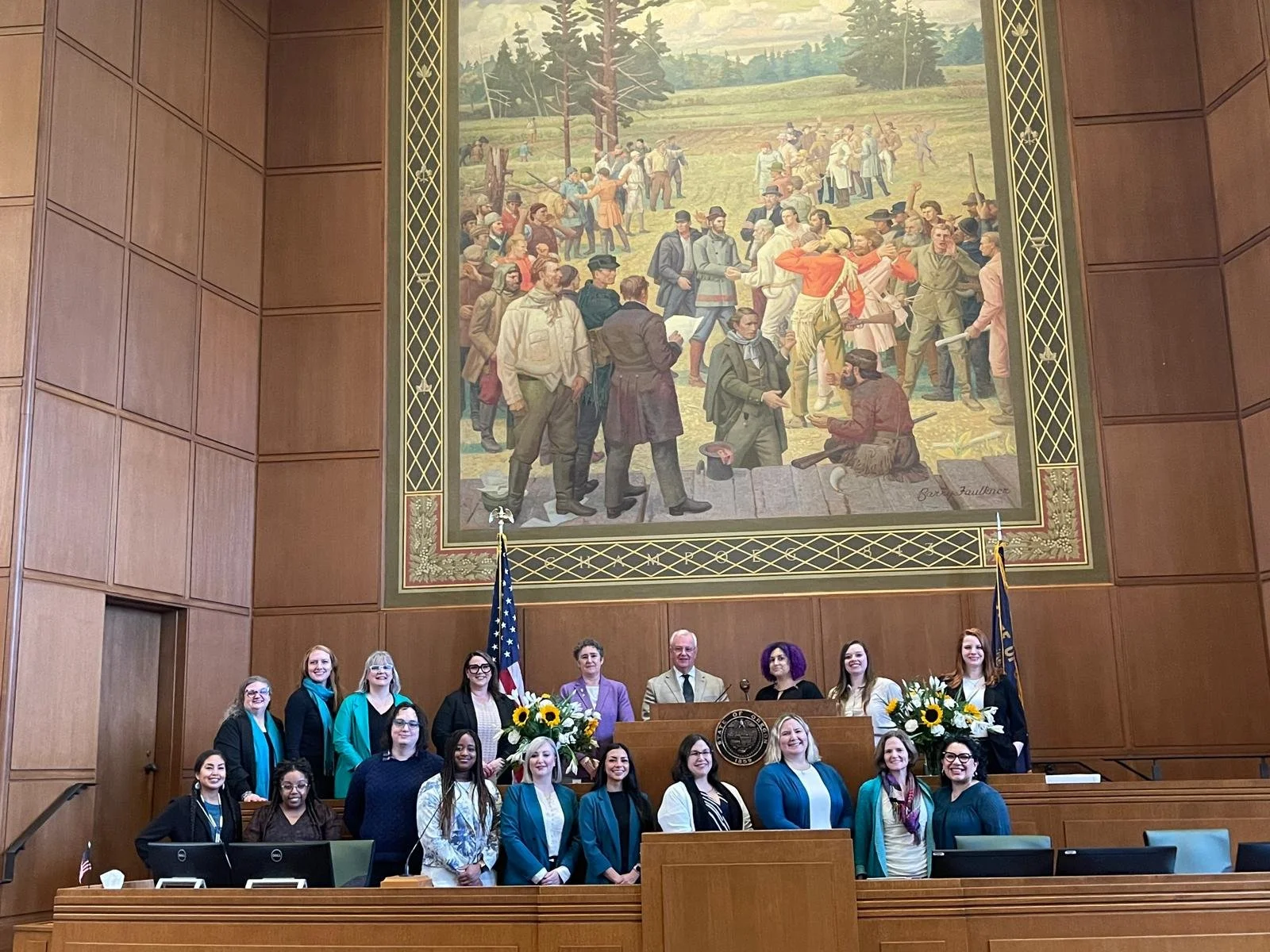 A group of diverse people standing and sitting in a wood-paneled legislative chamber, with a large historical mural on the wall behind them depicting a historical scene, two flags, and a podium with a seal, flowers, and a gavel.