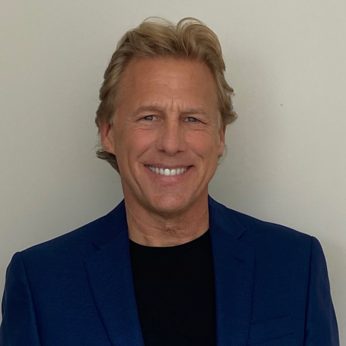 A man with blond hair and blue eyes smiling, wearing a dark blue blazer and a black shirt, standing against a plain light-colored wall.