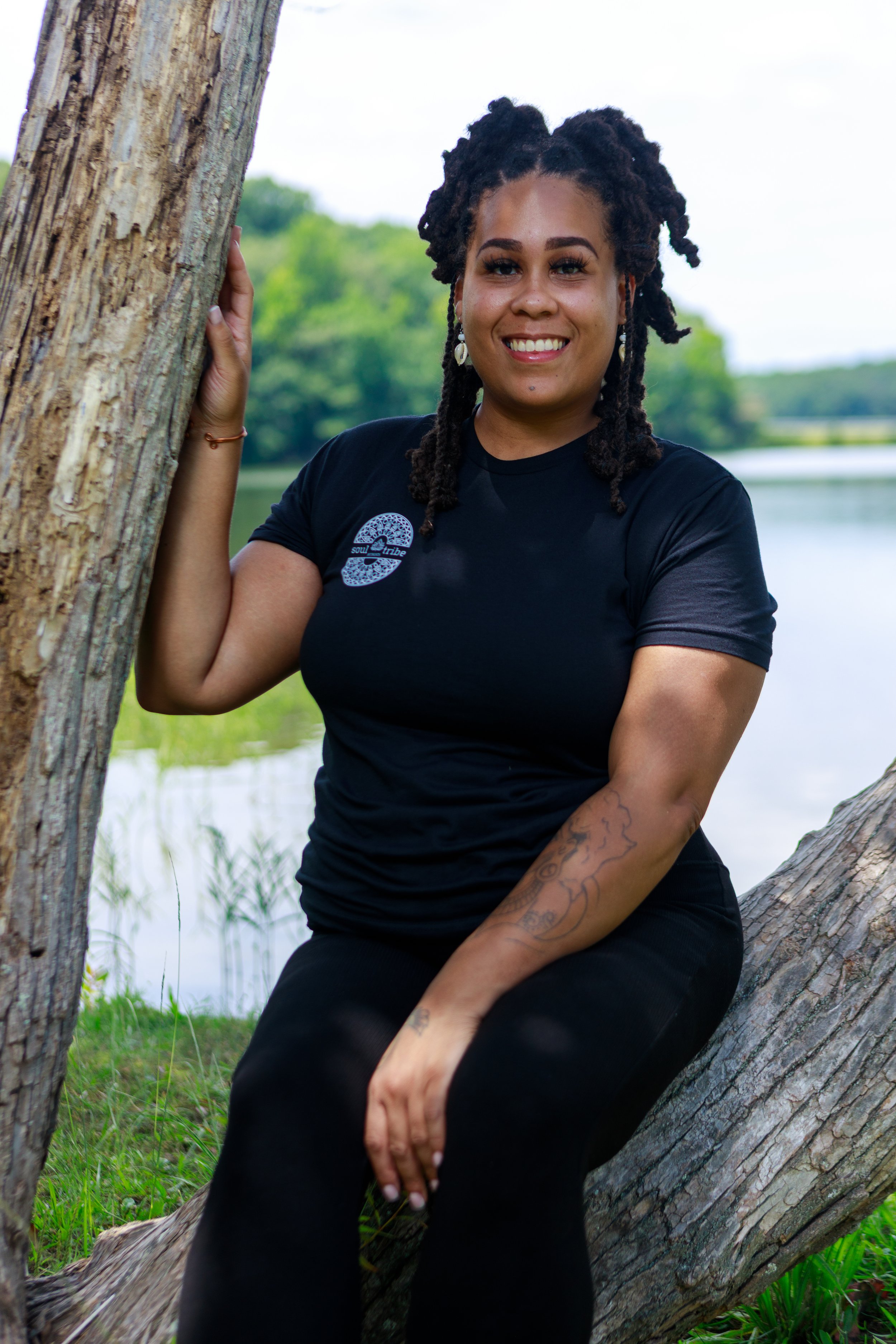 A woman with dark curly hair, wearing earrings and a black t-shirt, sitting by a tree near a body of water, smiling at the camera.