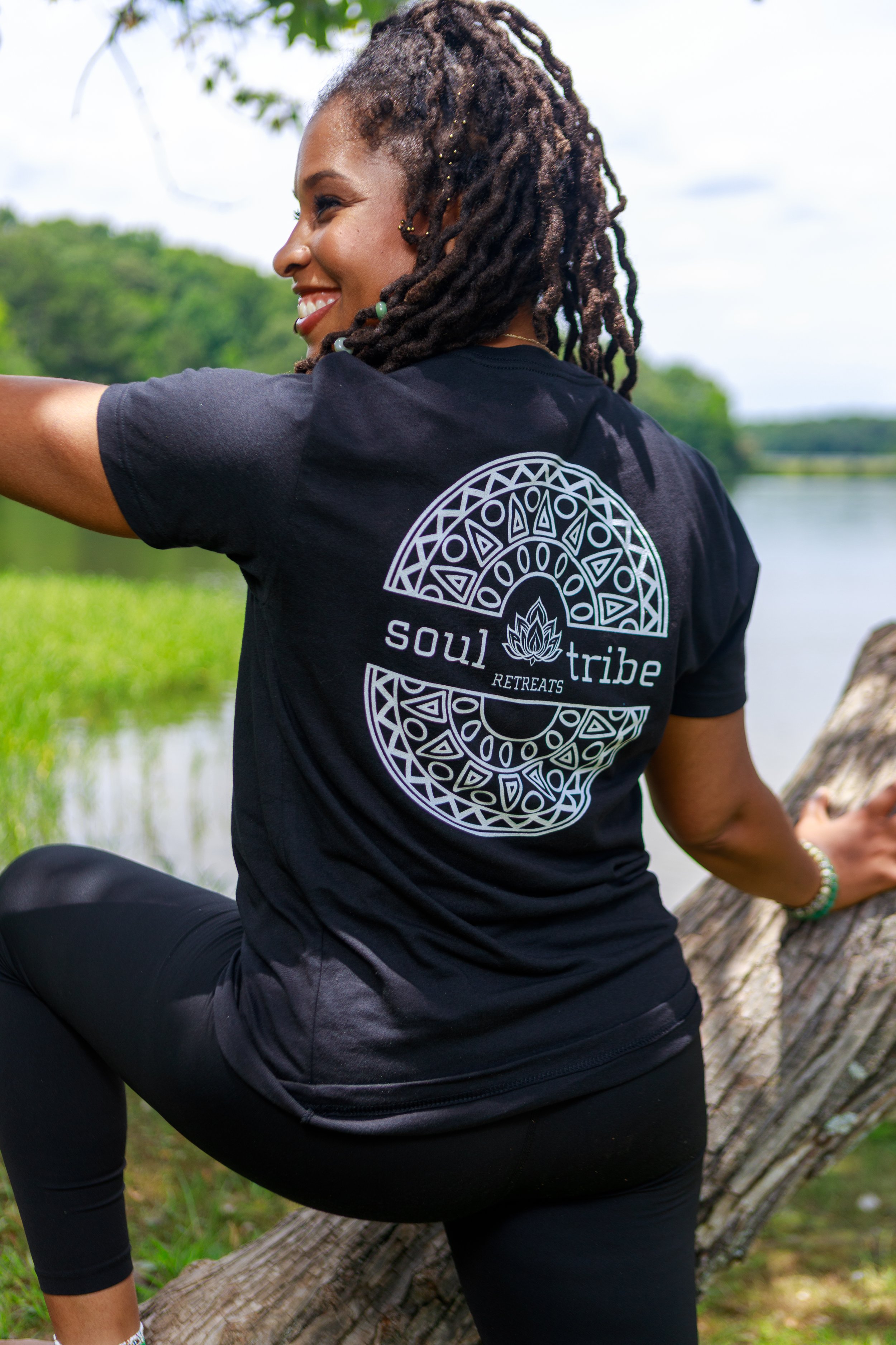 A woman with braided hair smiling while sitting on a fallen tree by a body of water, wearing a black t-shirt that has a logo with geometric patterns and the words 'soul tribe' and 'retreats'.