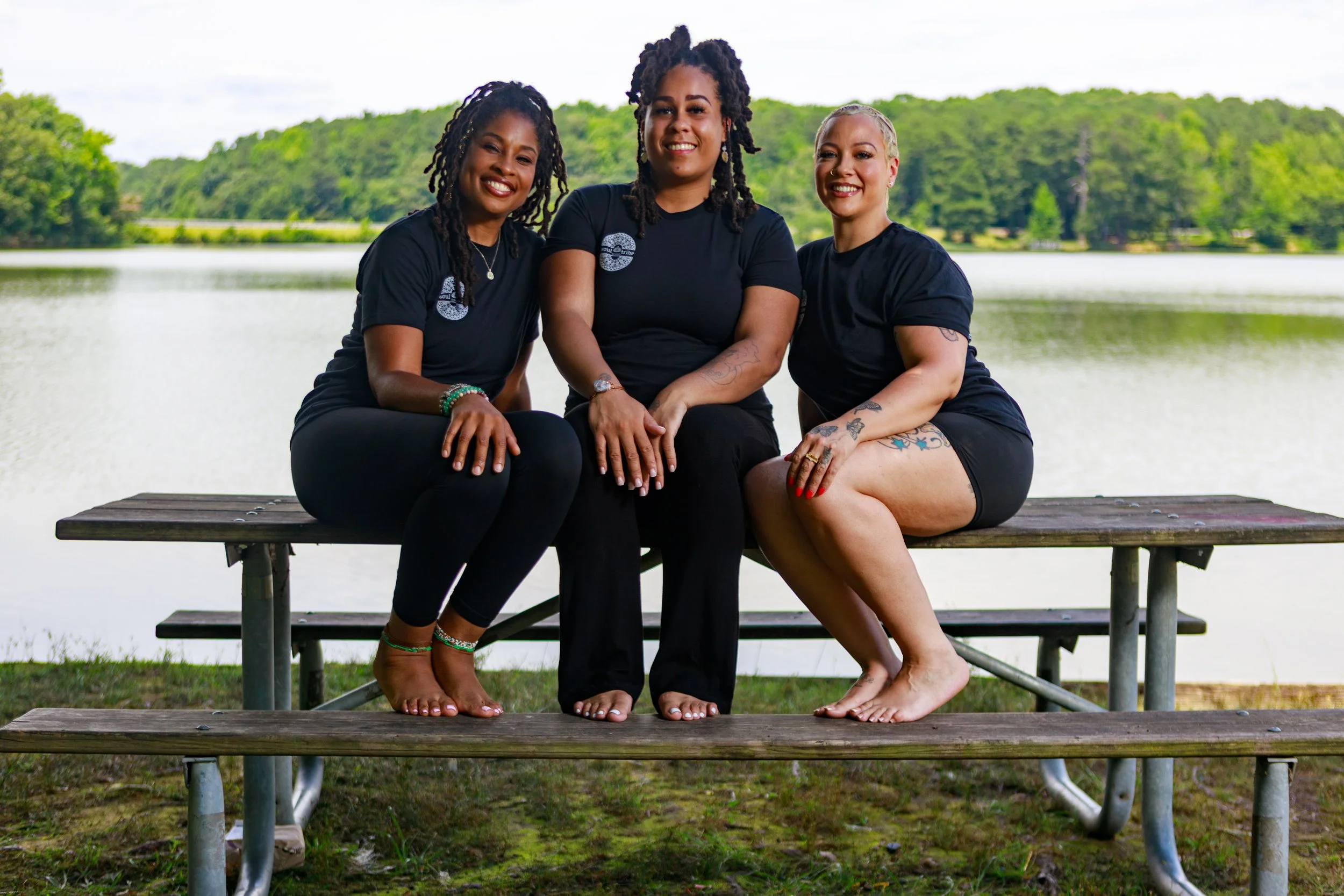Three women sitting on a wooden picnic table by a lake with green trees in the background, smiling at the camera.