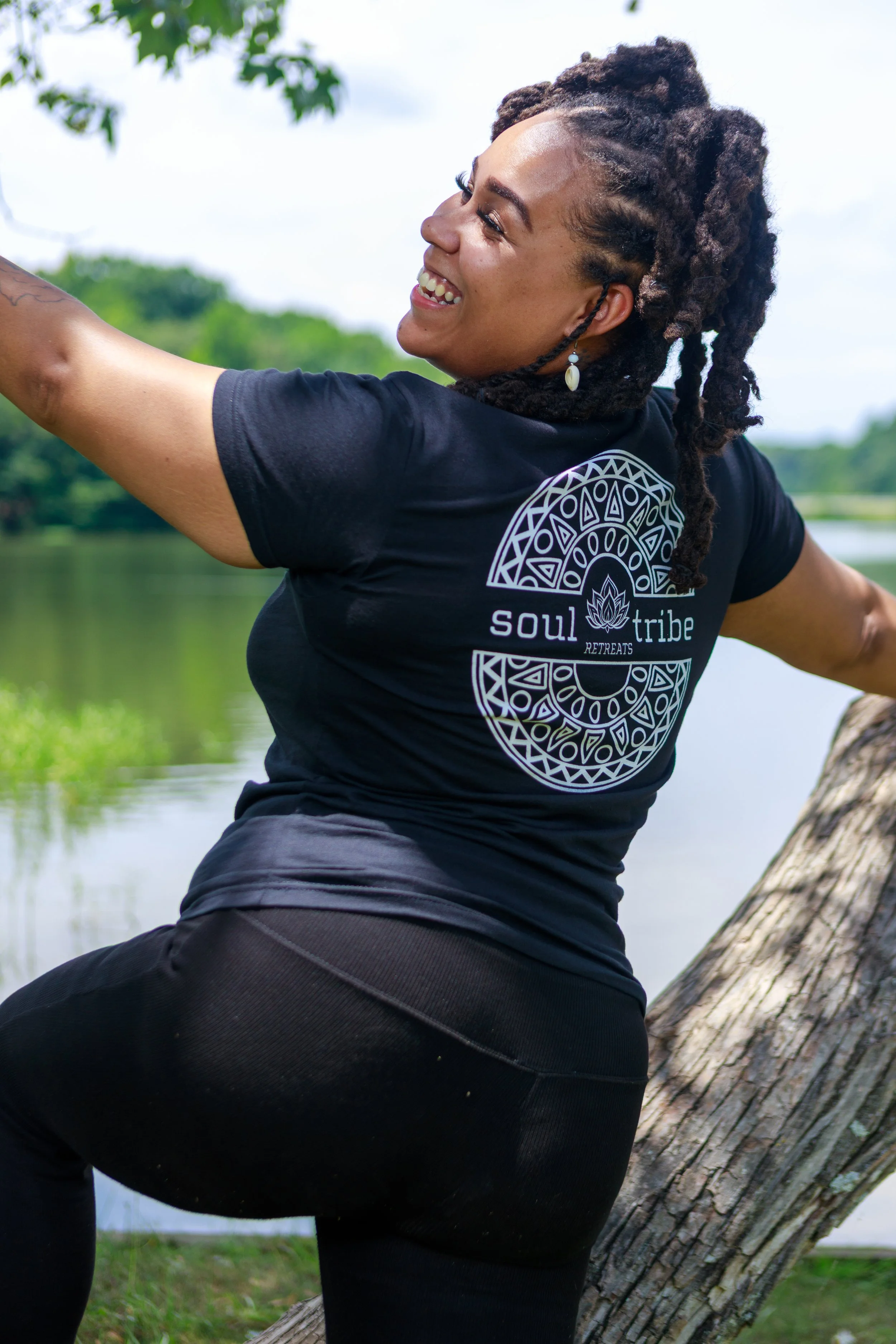 A smiling woman in a black T-shirt with a tribal design on the back and wearing shell earrings, enjoying outdoors near a body of water and trees, with her arms raised, leaning against a large tree trunk.