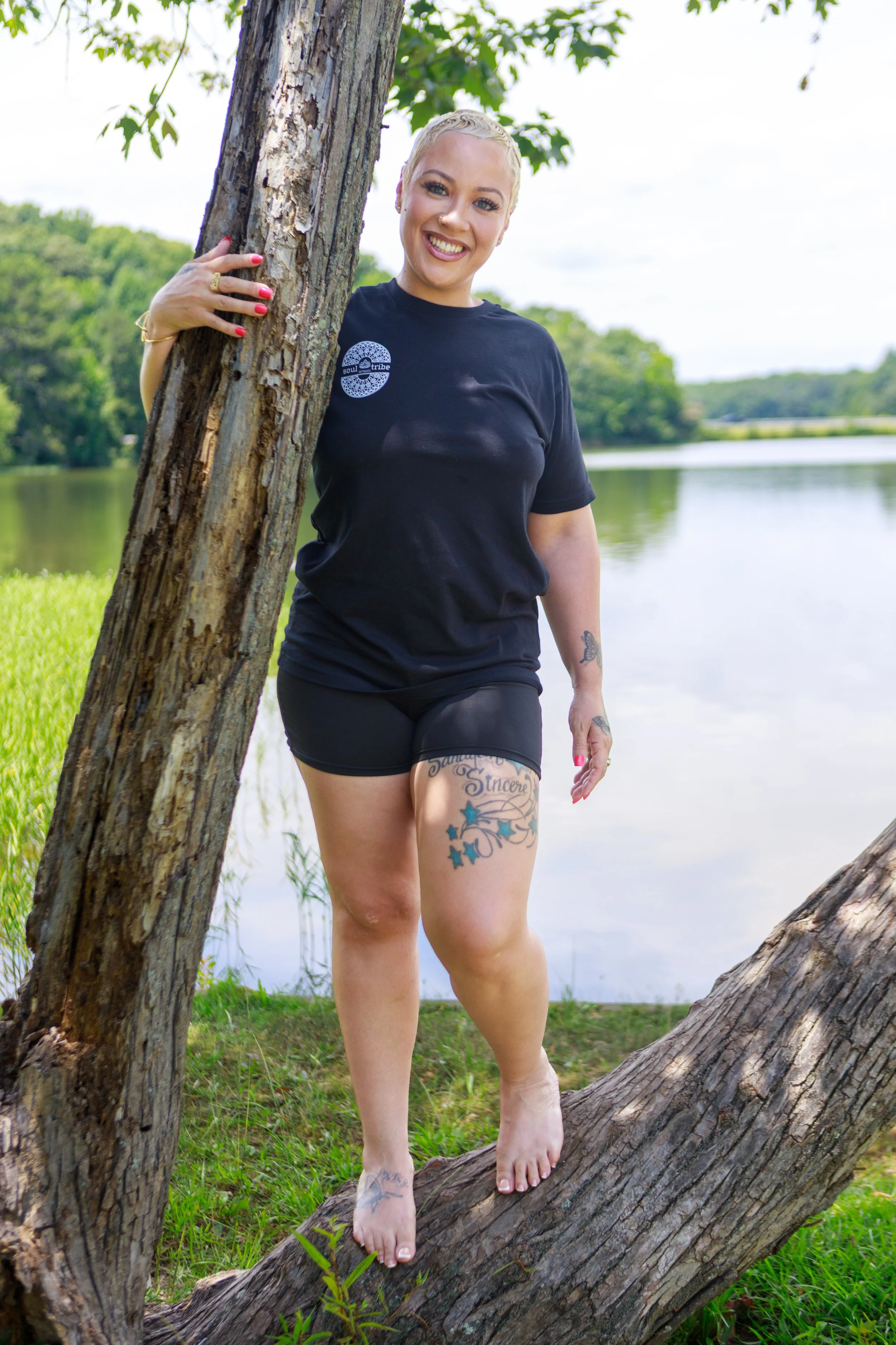 A woman with short blonde hair, tattoos on her thighs, and wearing a black t-shirt standing outdoors by a lake, holding onto a tree branch and smiling.