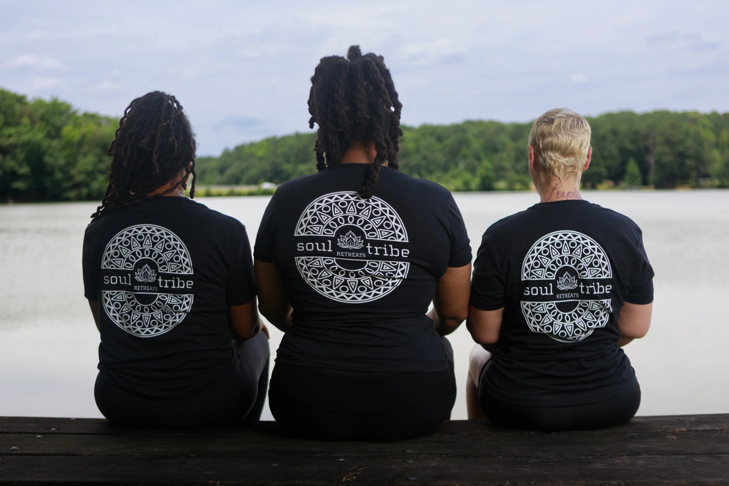 Three individuals sitting on a wooden surface outdoors, facing away, wearing matching black t-shirts with a circular design and the words "soul tribe RETREATS" on the back, overlooking a body of water with trees in the background.