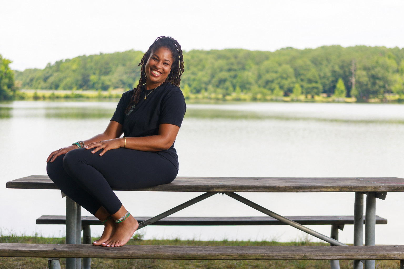 A woman in black clothes sitting cross-legged on a wooden picnic table near a lake, smiling, with green trees and water in the background.