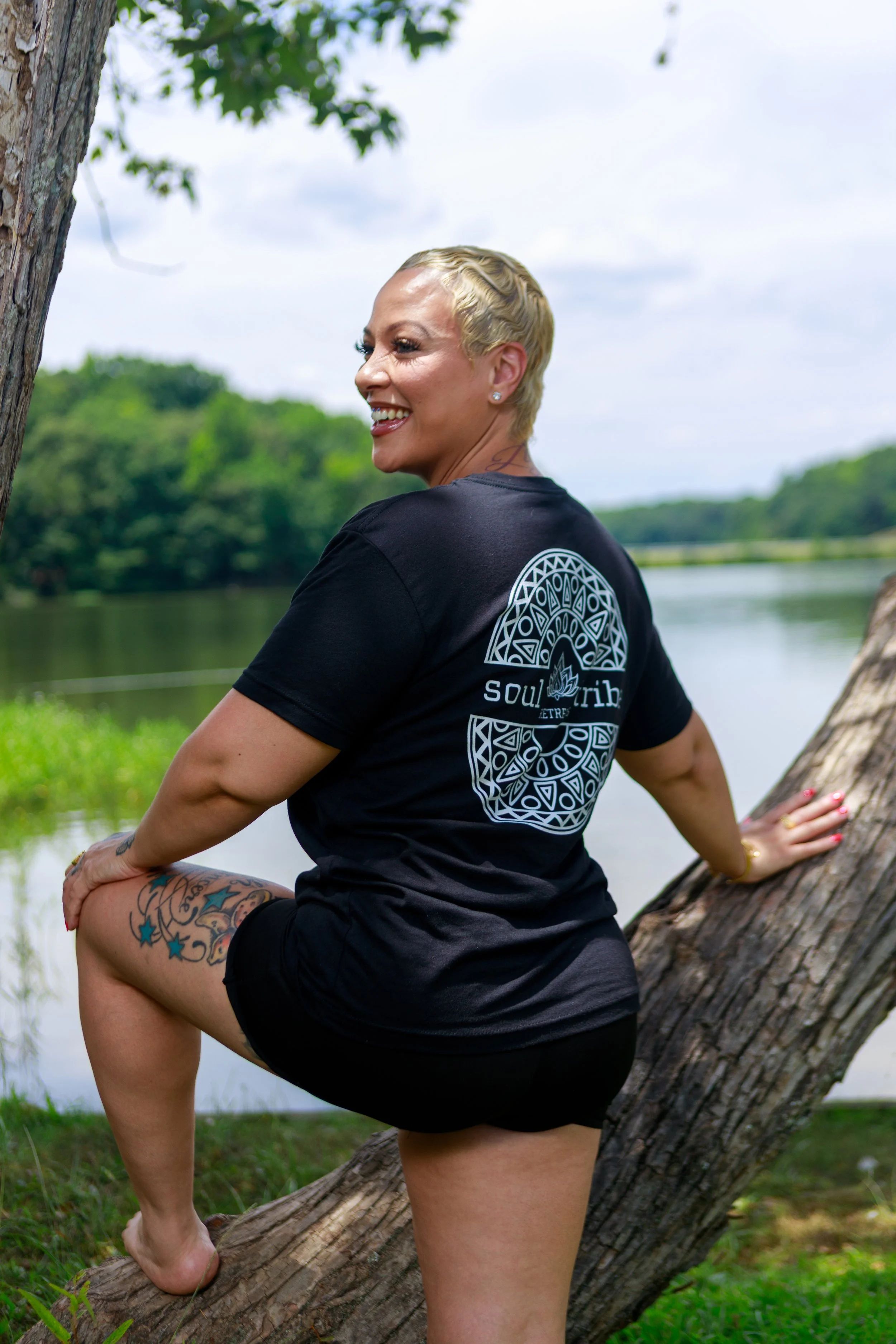 A woman with short blonde hair practicing yoga outdoors on a tree branch near a body of water, wearing a black t-shirt with a white design on the back and black shorts, smiling and looking to the side.