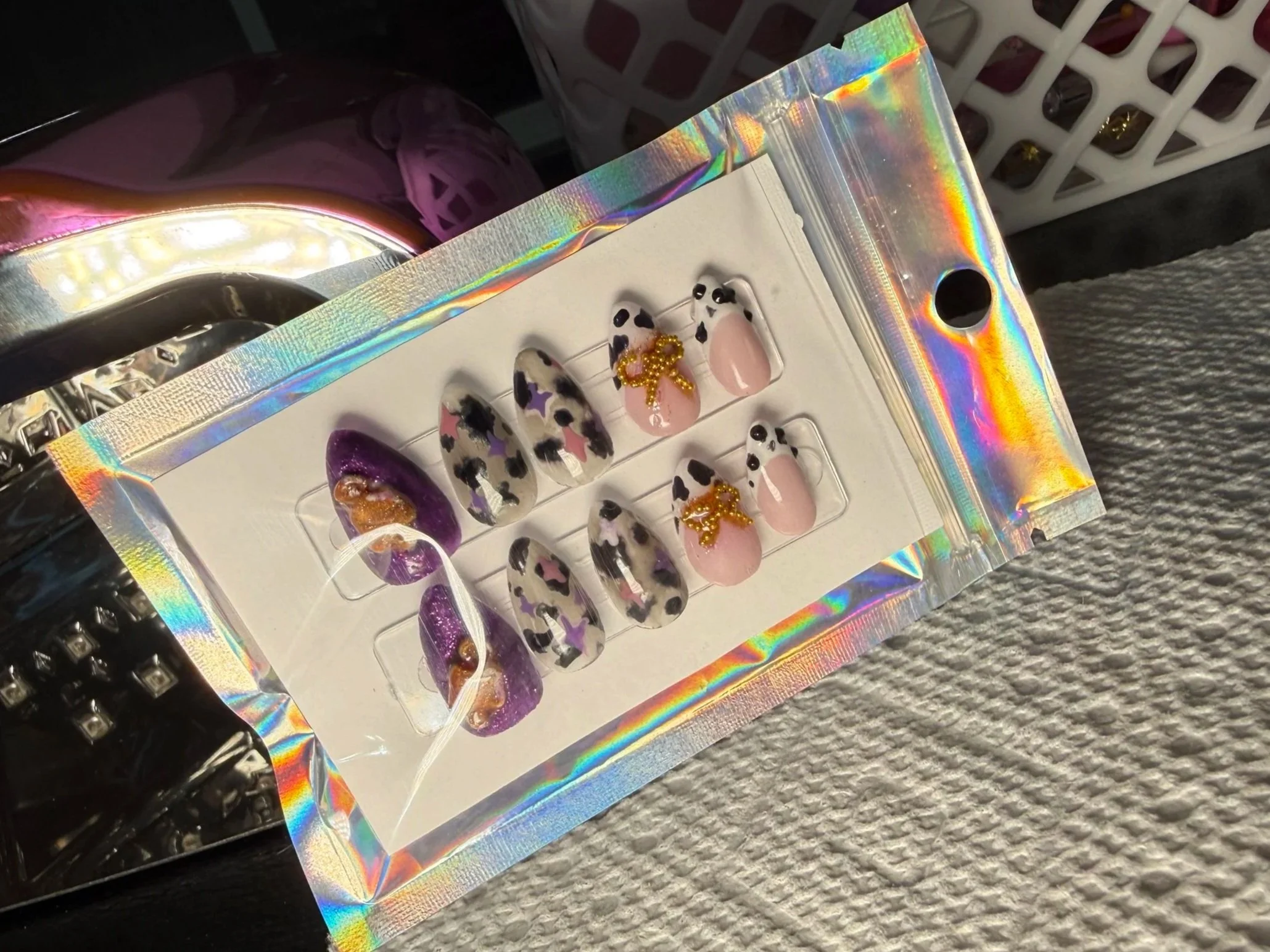 Holographic display case with assorted false nails decorated with leopard print, butterflies, and bows, placed on a glittery surface.