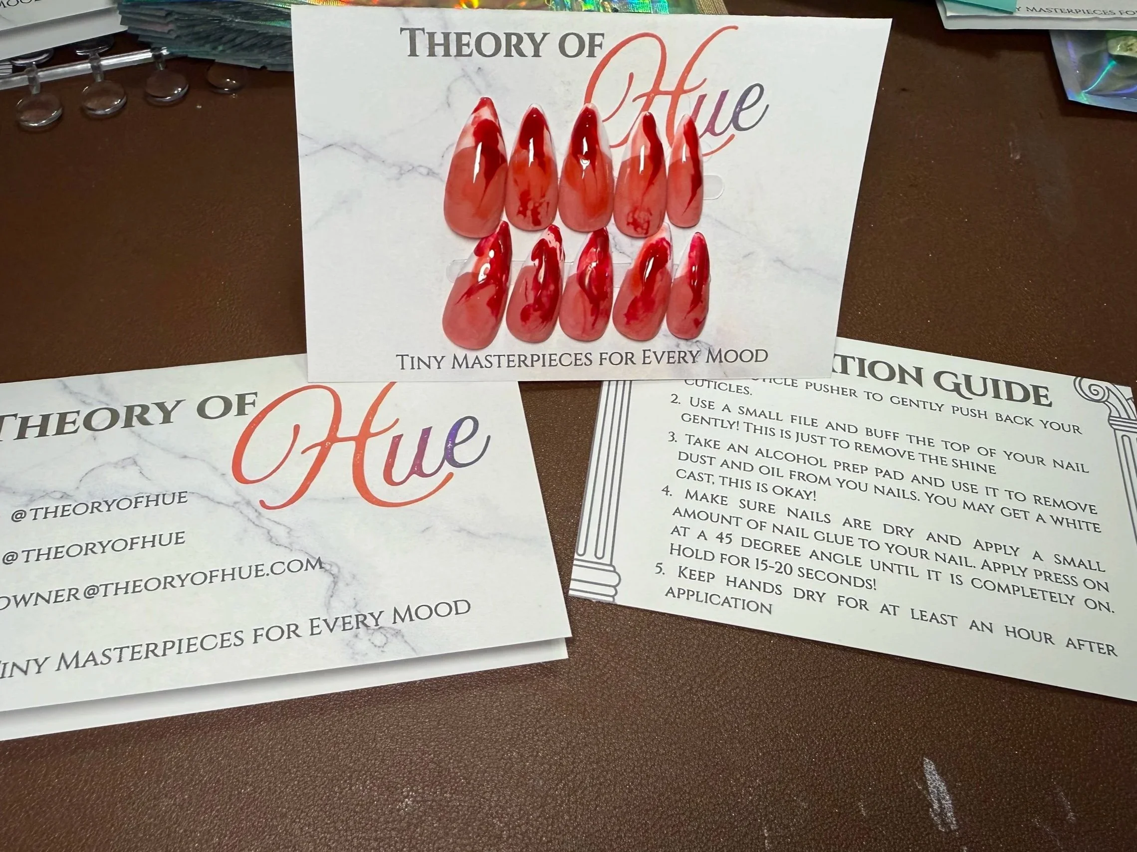Display card showing examples of red nail designs labeled "Theory of Hue," with the subtitle "Tiny Masterpieces for Every Mood". Surrounding papers include nail care instructions.