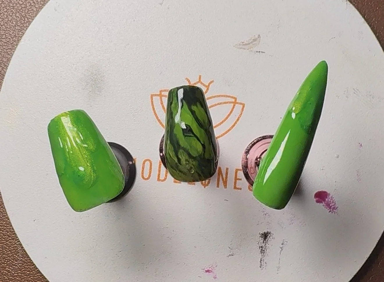 Three colorful, glossy nail polish swatches on a white circular surface with an orange logo and some pink and black paint smudges.