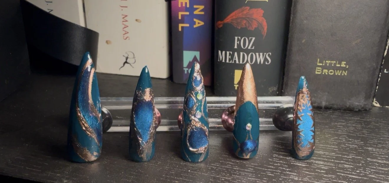 Five artfully painted hair curlers with blue, gold, and pink designs arranged on a black surface in front of a bookshelf.