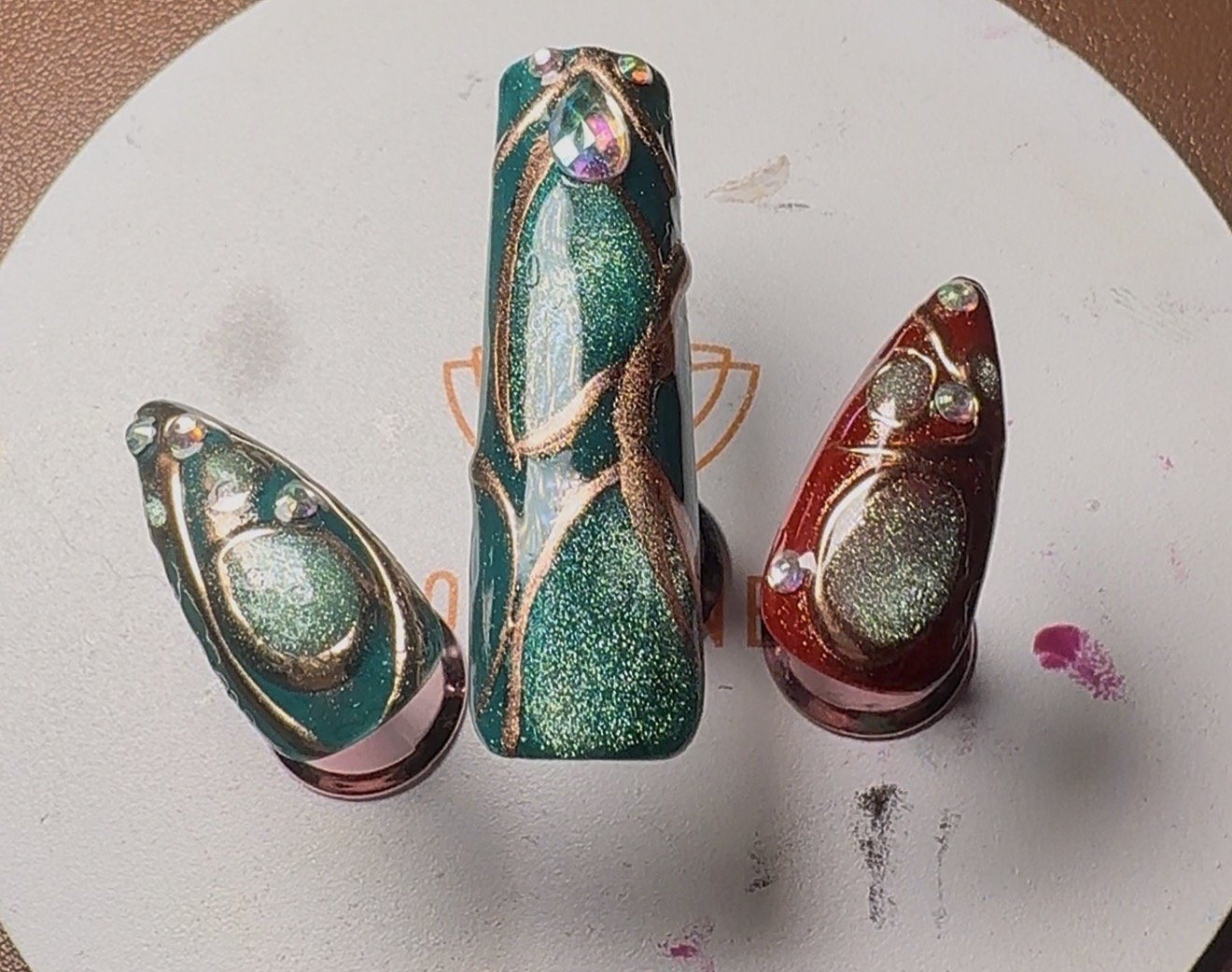 Close-up of three decorative glass nail art designs with gold swirls and embedded glitter, set on a textured surface with paint smudges.