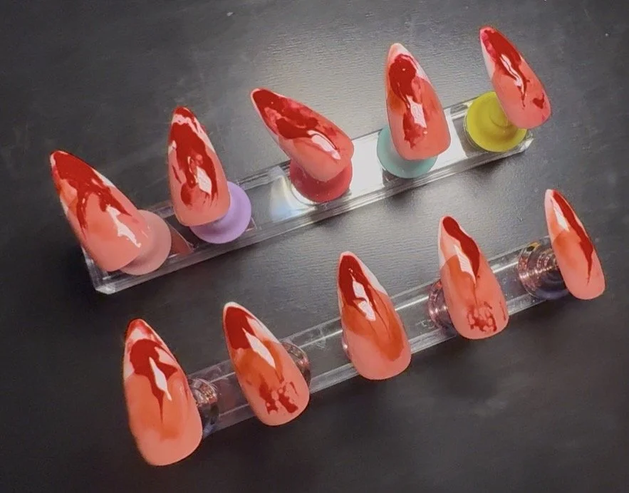 Two clear acrylic display stands with ten artificial nails painted in red and pink with a glossy finish, arranged on a dark surface.