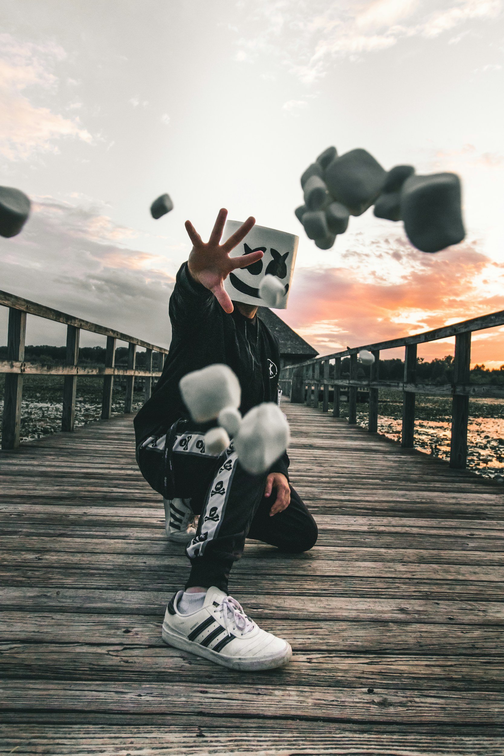 EDM DJ Marshmello standing on a wooden boardwalk, throwing white marshmallows directly toward the camera at sunset.