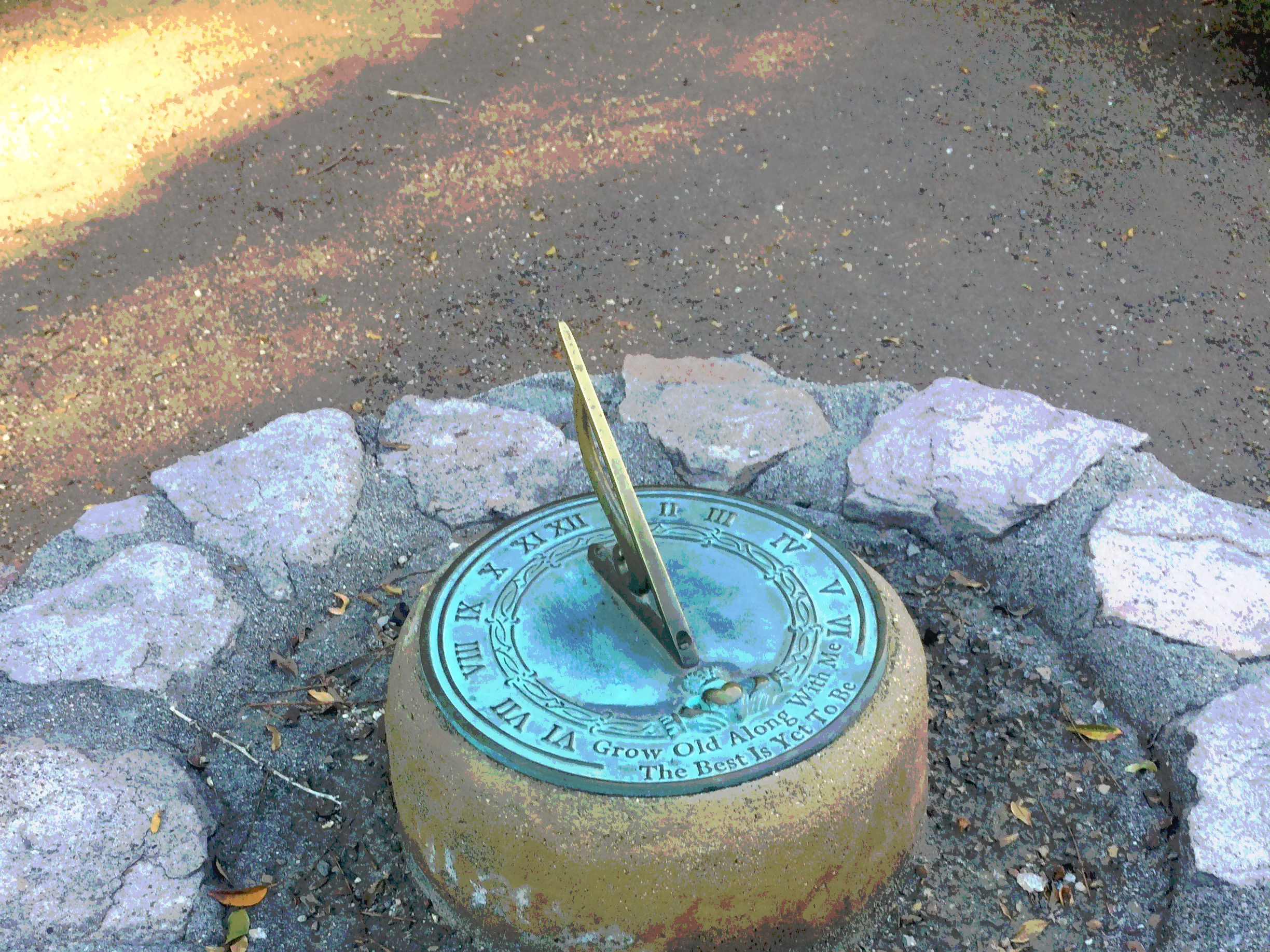 A sundial surrounded by rocks on a dirt path, with a yellowish-green grassy area in the background.