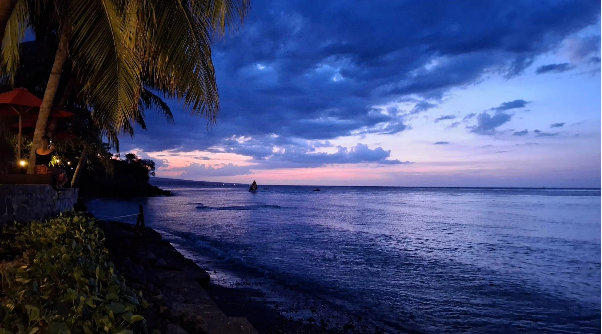 Blue-hour ocean view in Bali with palm trees, calm water, and a small sailboat on the horizon.