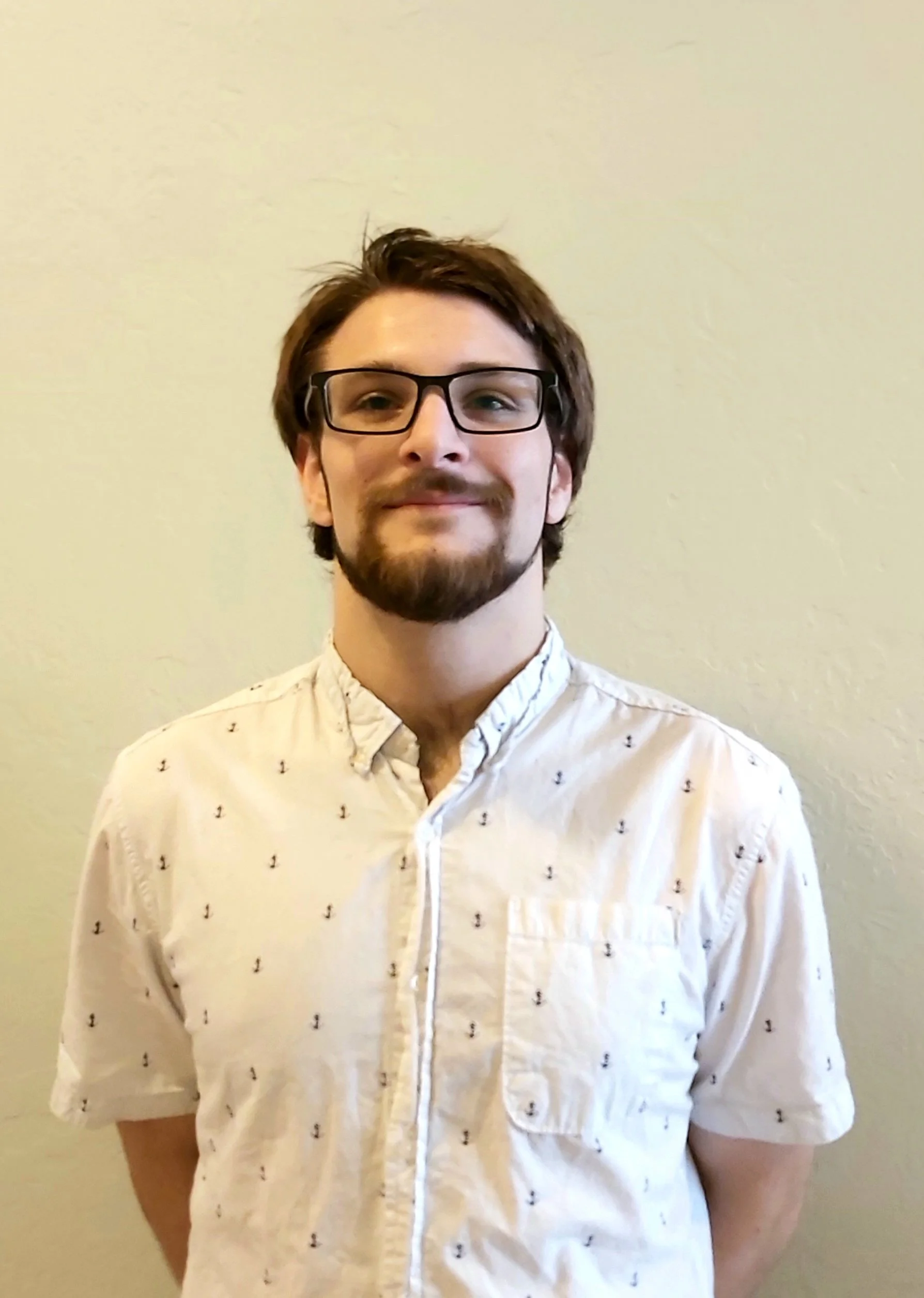 Portrait of a young man with glasses, brown hair, and a beard, wearing a cream-colored shirt with small black anchor patterns, standing against a light-colored wall.