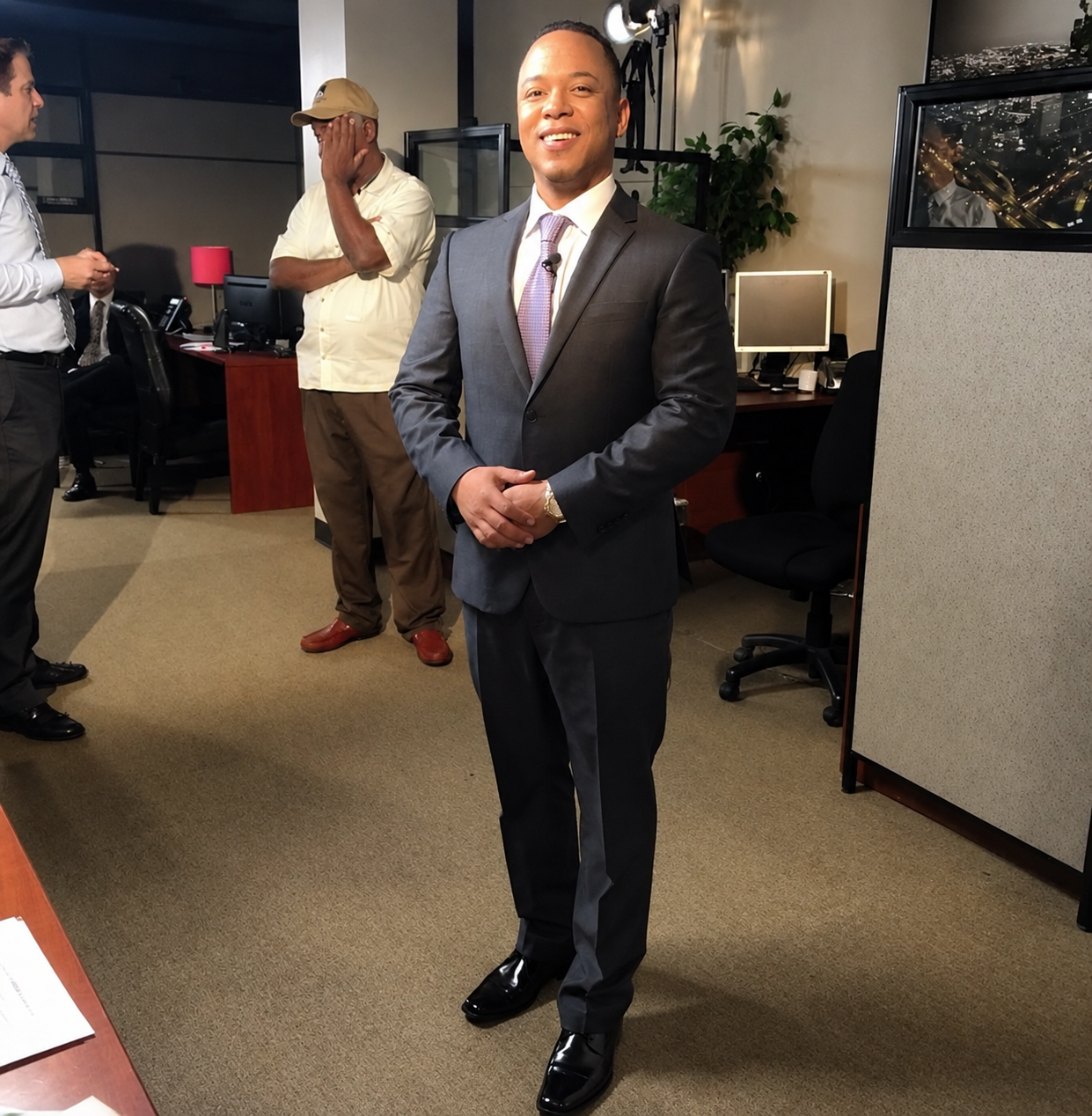 Man in a gray suit and purple tie standing confidently in an office, with three other men in the background. One man with a beige cap covering his face, another in a white shirt, and a third in a white shirt and tie, engaged in conversation.