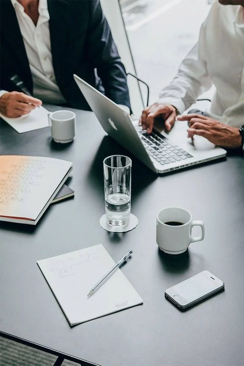 Two people sitting at a table with laptops, notebooks, pens, coffee cups, a glass of water, and a smartphone.