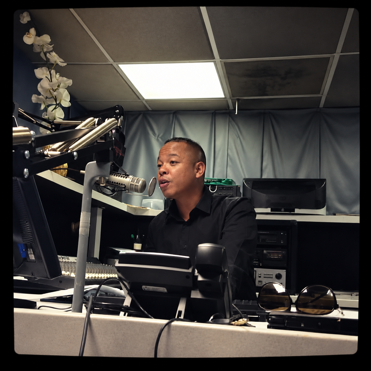 A man speaking into a microphone in a radio station studio, surrounded by radio equipment, with gray curtains and a ceiling with fluorescent lighting.