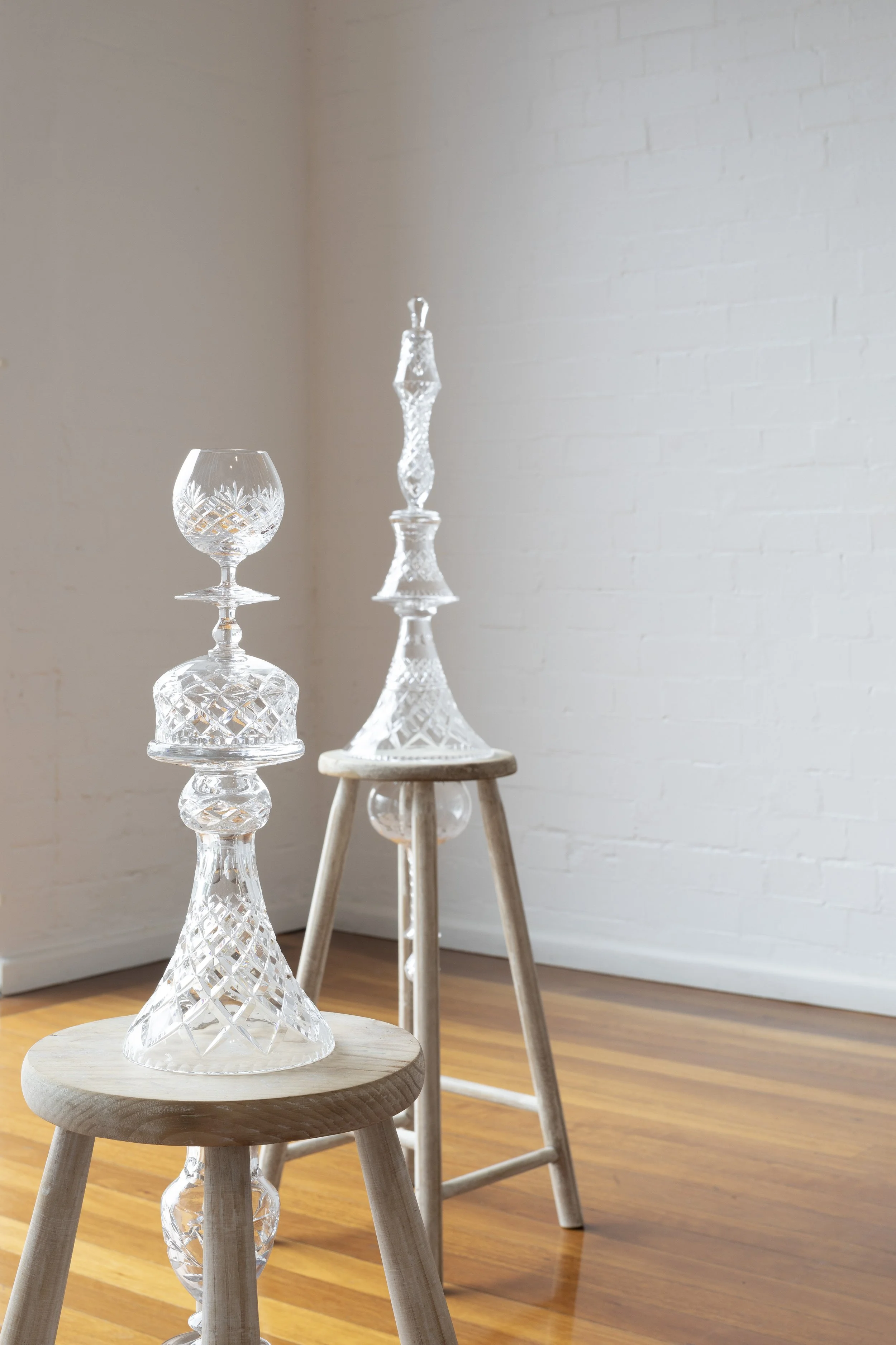 Three glass decanters with intricate cut patterns on small wooden stools in a room with white brick wall and hardwood floors.