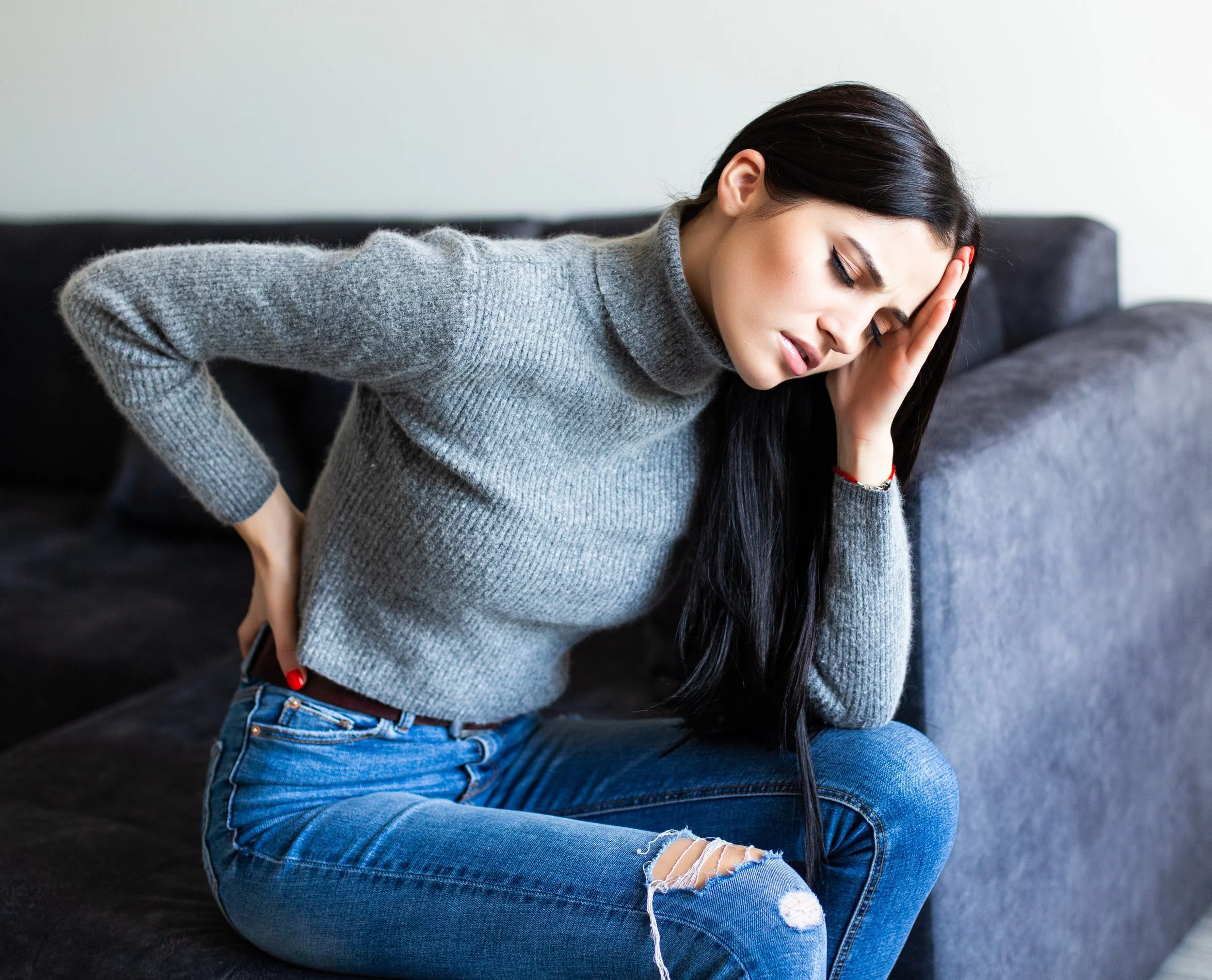 A young woman with long dark hair wearing a gray turtleneck sweater and ripped blue jeans sitting on a dark sofa with her eyes closed and hand on her head, appearing to be in pain or discomfort.