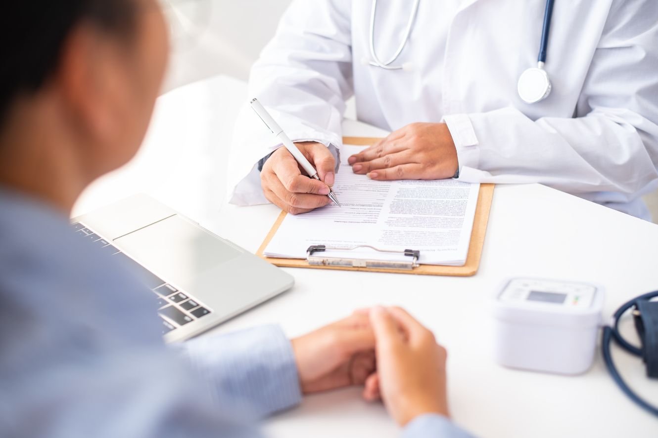 Doctor taking notes on a clipboard while consulting with a patient. Medical equipment on the table.