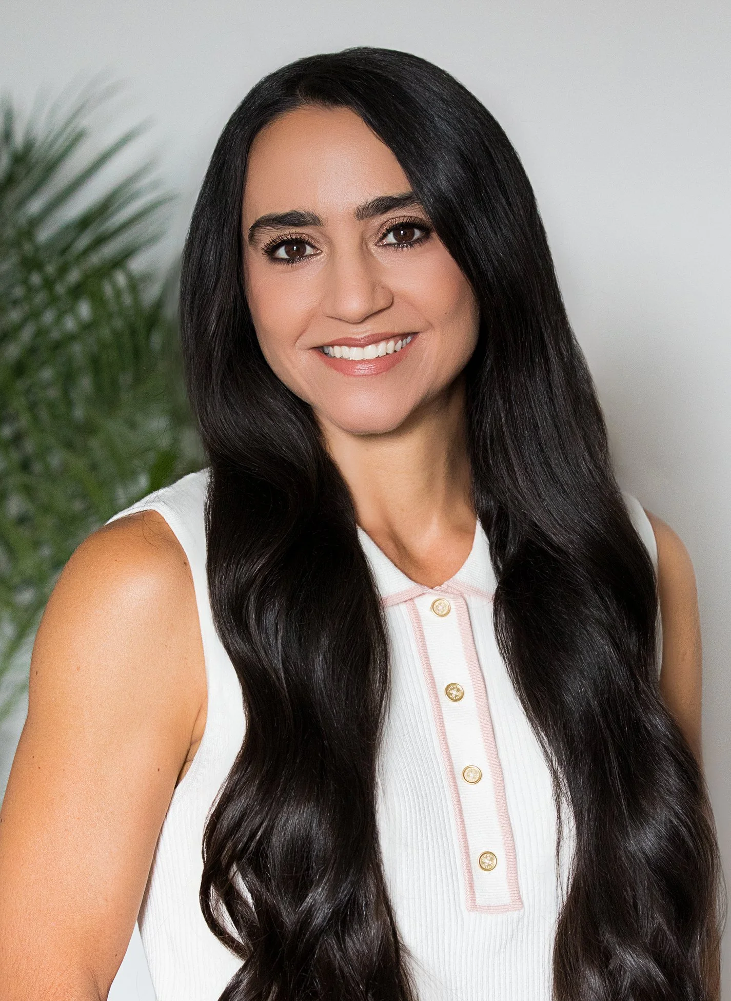 A woman with long dark hair, smiling, wearing a white sleeveless top with pink buttons and a pink outline, in front of a blurred background with green plants and a white wall.