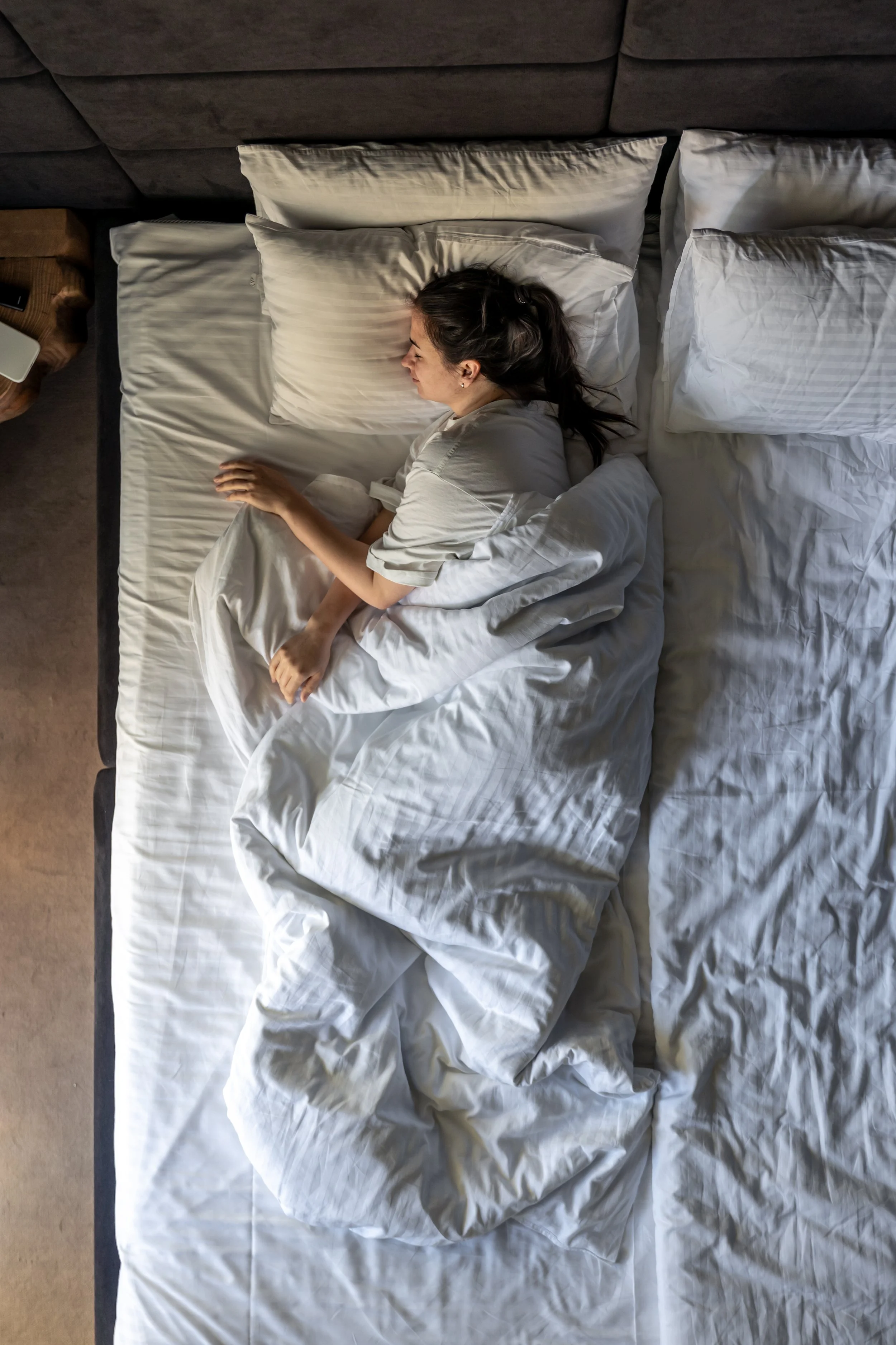 A woman with long dark hair sleeping on her side in a bed with white linens, next to a pillow, with a grey headboard and a wooden nightstand with objects beside it.
