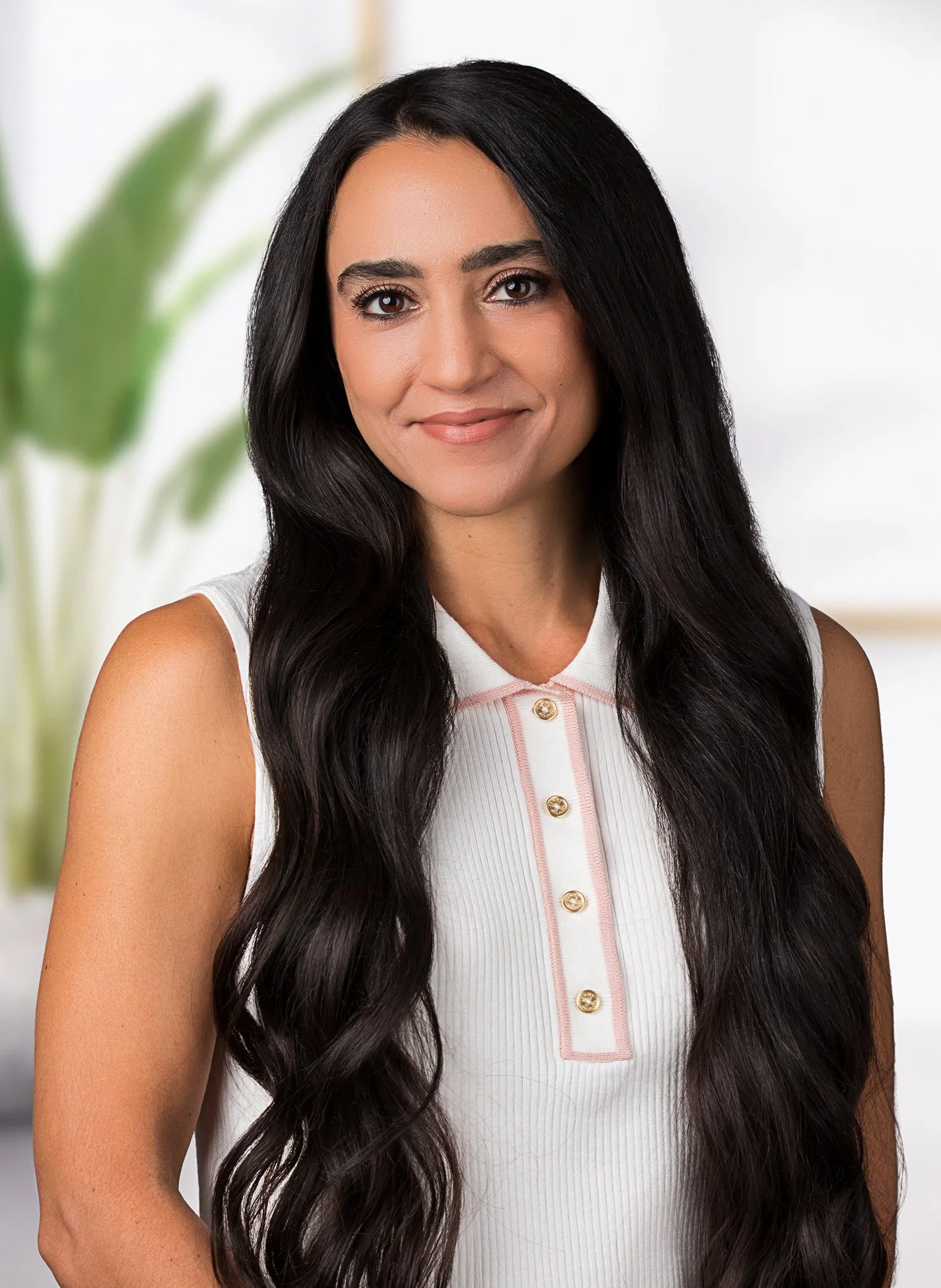 A woman with long, dark wavy hair and brown eyes smiling at the camera, wearing a sleeveless white top with pink trim and gold buttons, with a blurred green plant in the background.