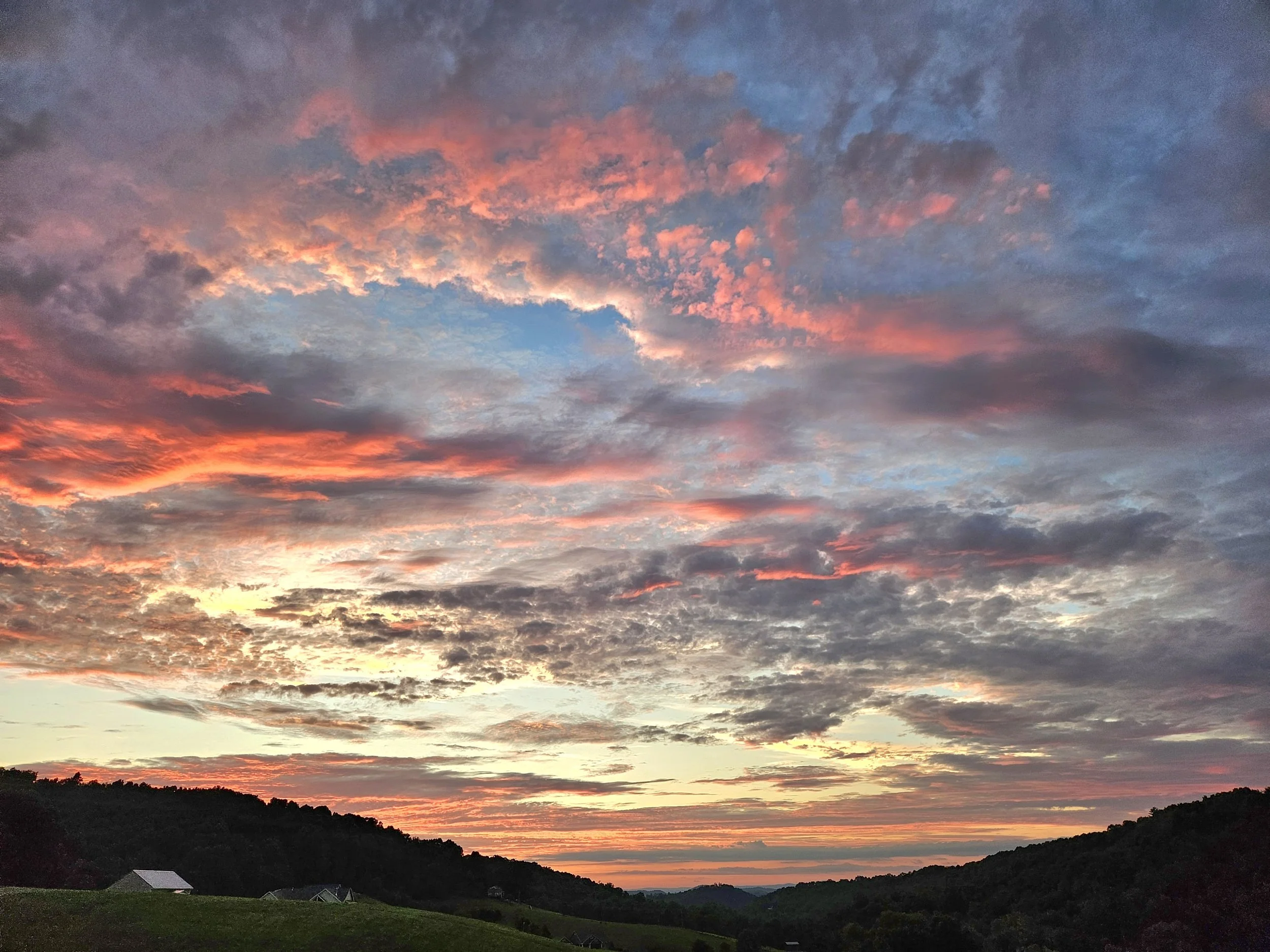 A scenic view of a colorful sunset with clouds in the sky, illuminating with shades of pink, orange, and blue, over a landscape of rolling hills and a few small houses.
