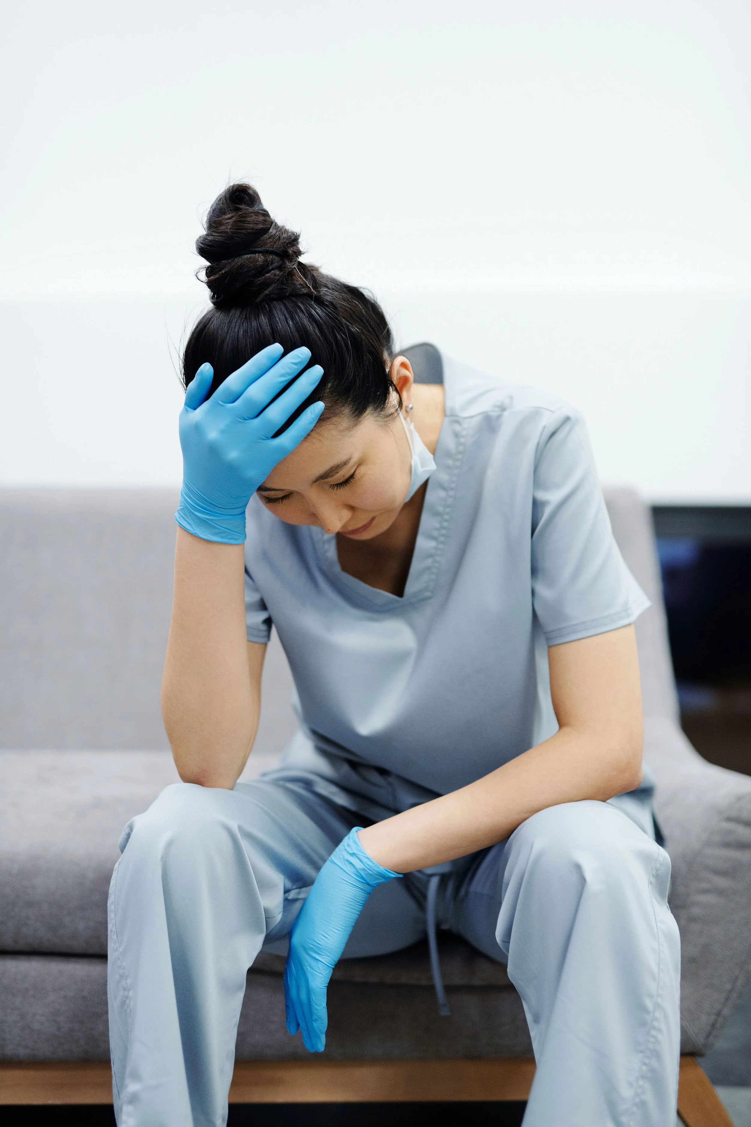 A woman in medical scrubs and blue gloves sitting on a bench with her head in her hand, looking down with a distressed expression.