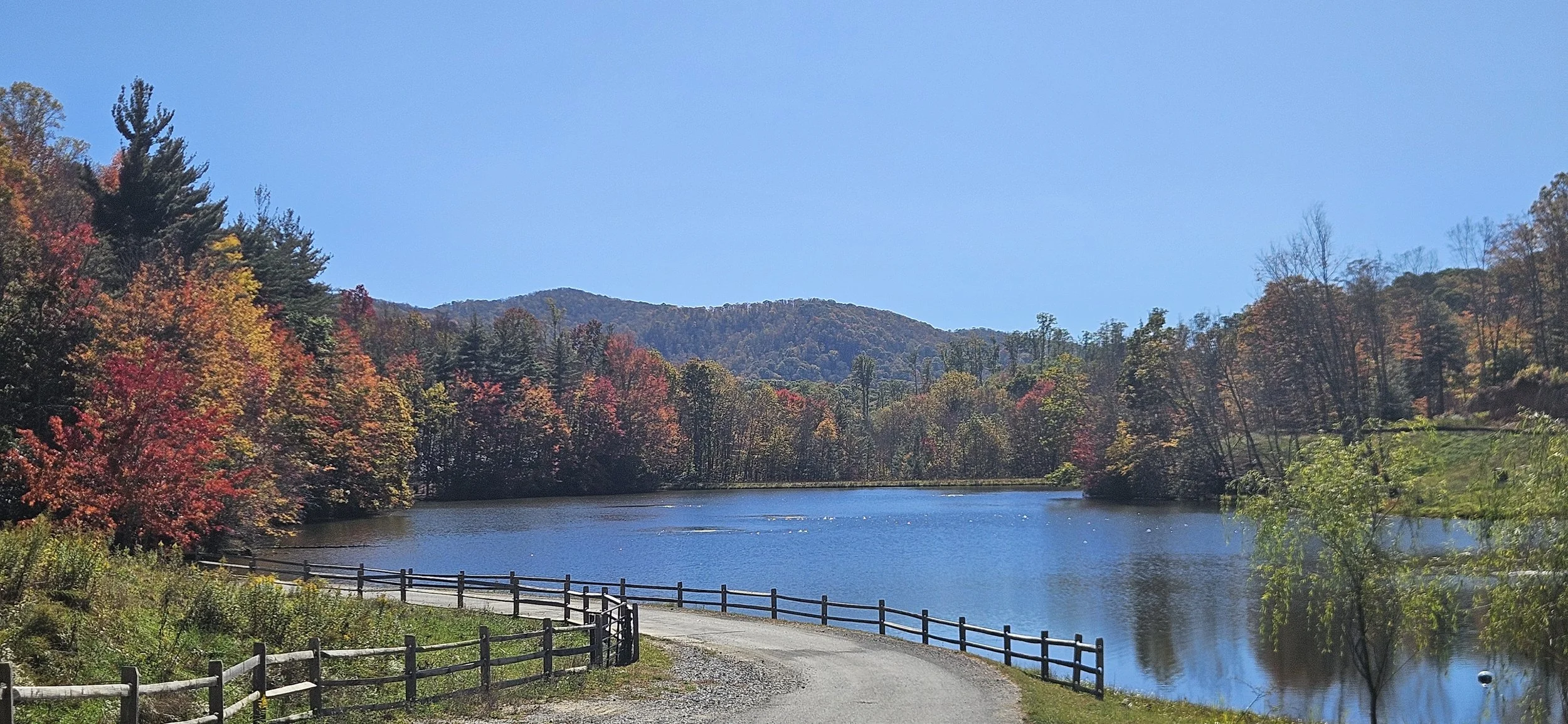 A scenic landscape featuring a calm lake surrounded by colorful autumn trees with red, orange, and yellow leaves, against a backdrop of rolling hills and a clear blue sky. A gravel road with a wooden fence runs along the lake's edge.