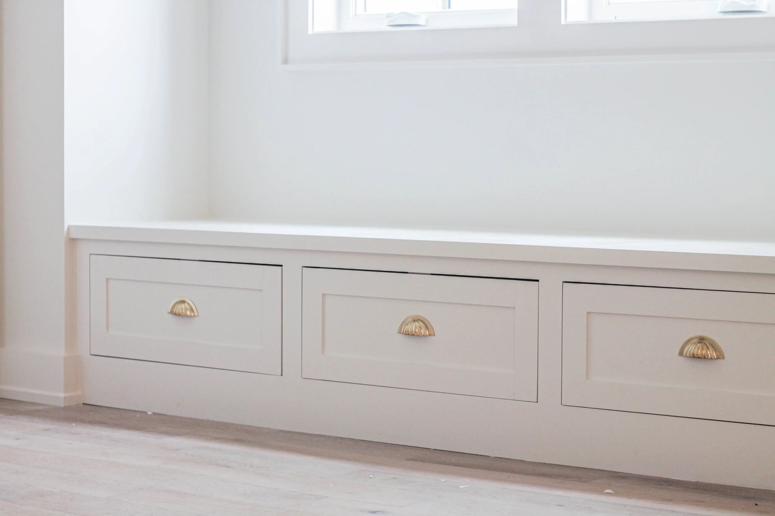 White built-in cabinet with drawers and rounded brass handles in a room with light-colored walls and floor.