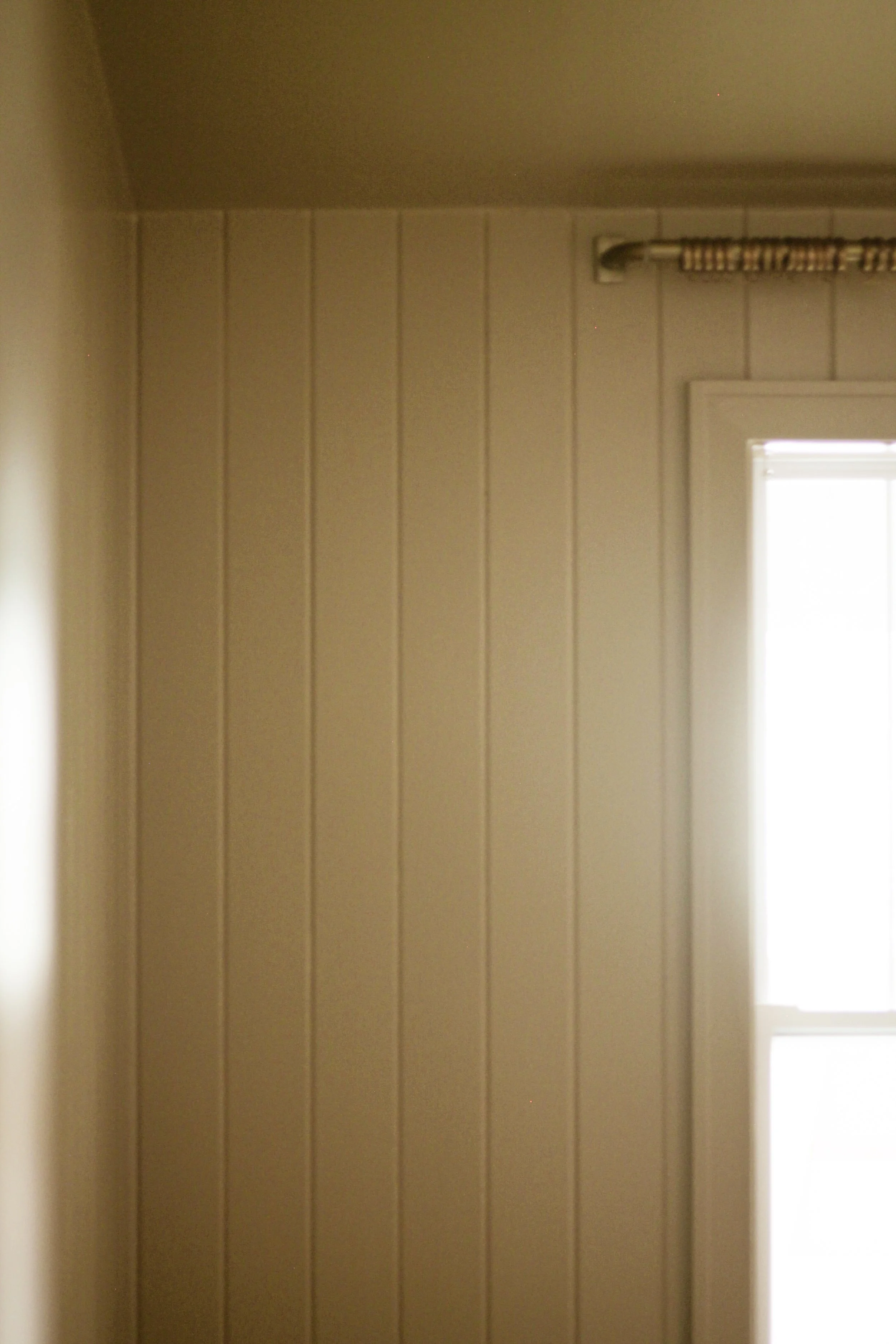 A corner of a room with beige wood-paneled walls, a window in the lower right corner with sunlight coming through, and a curtain rod above the window.
