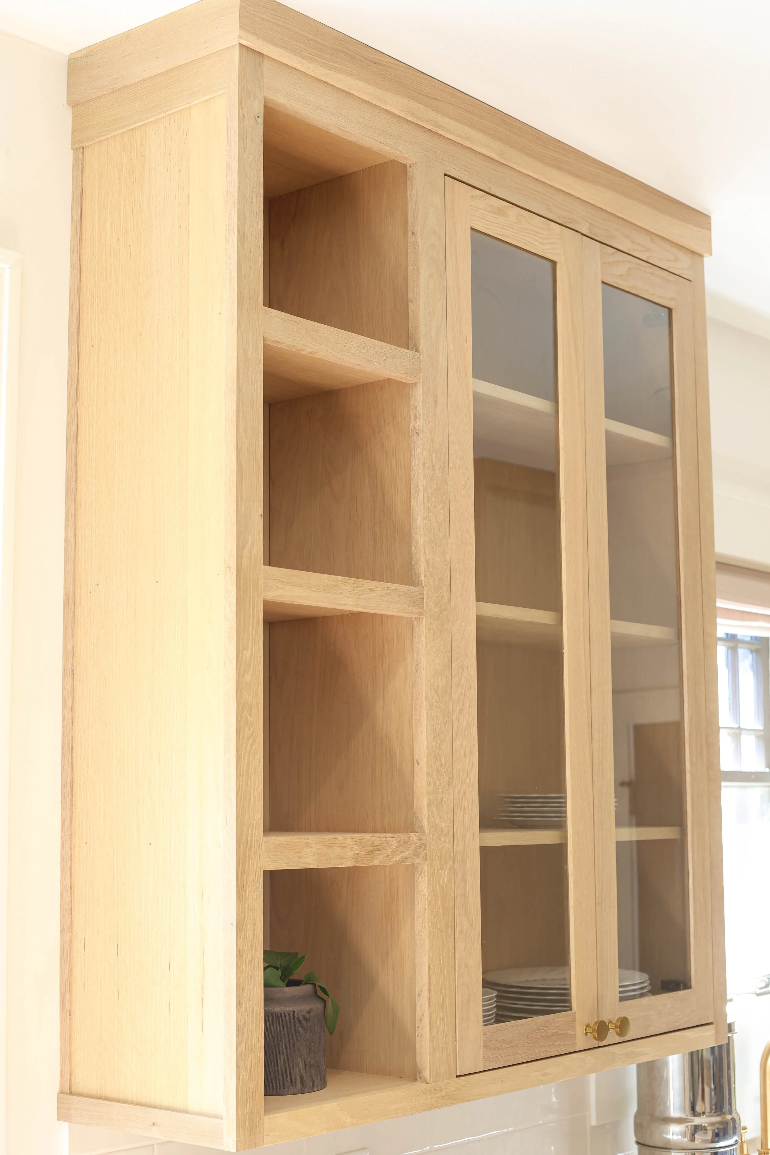 Light wood kitchen cabinet with glass doors and brass knobs, open shelves with plates and a potted plant inside, next to a light-colored wall and window.