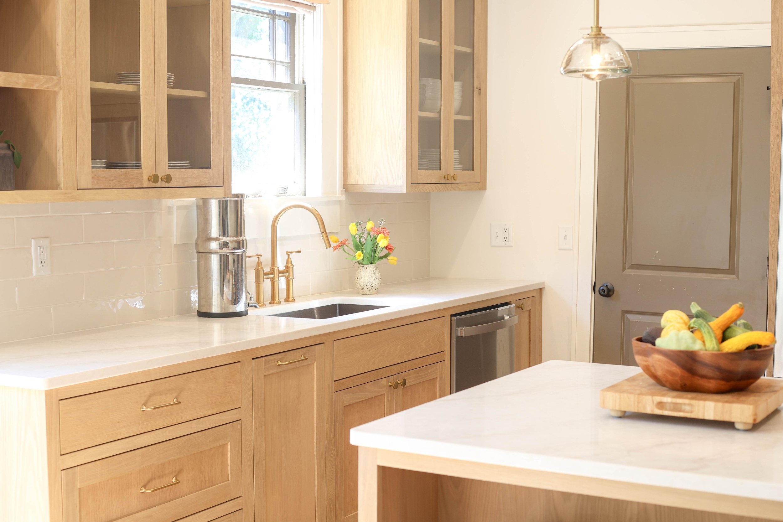 A bright kitchen with wooden cabinets, a window, a sink with a brass faucet, a bouquet of flowers, and a wooden fruit bowl on an island.