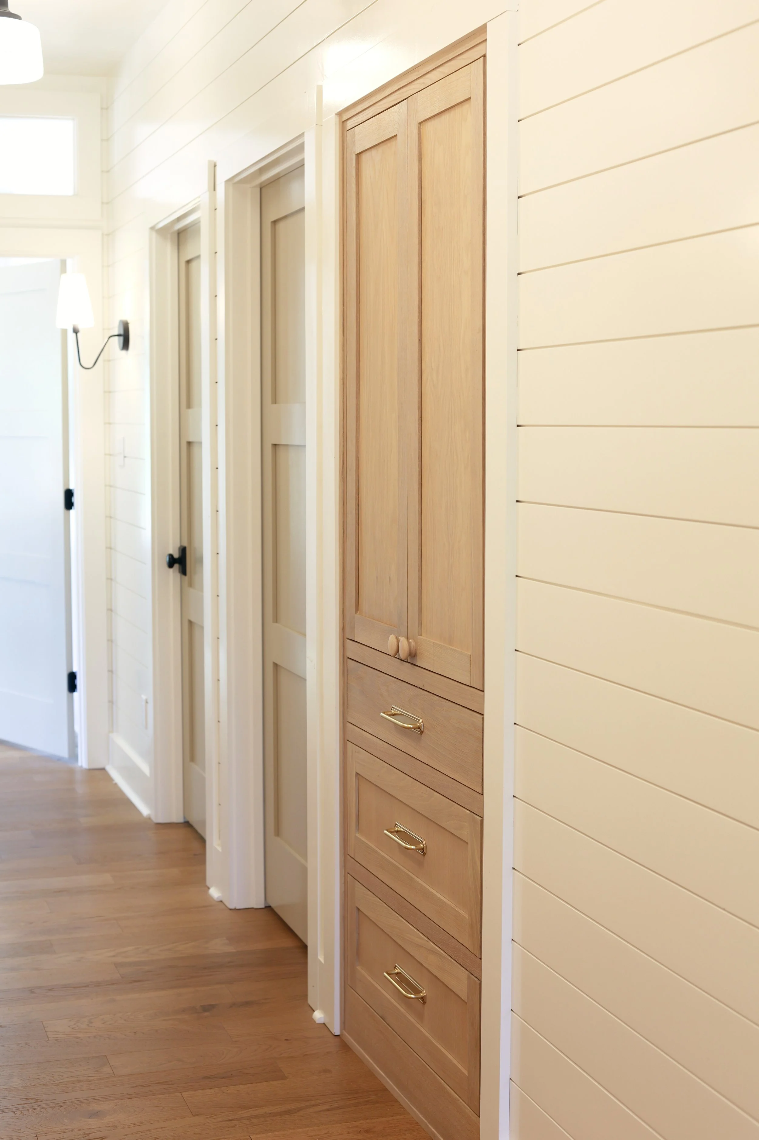 Hallway with white shiplap walls, hardwood flooring, and a wooden cabinet with gold handles mounted on the wall.