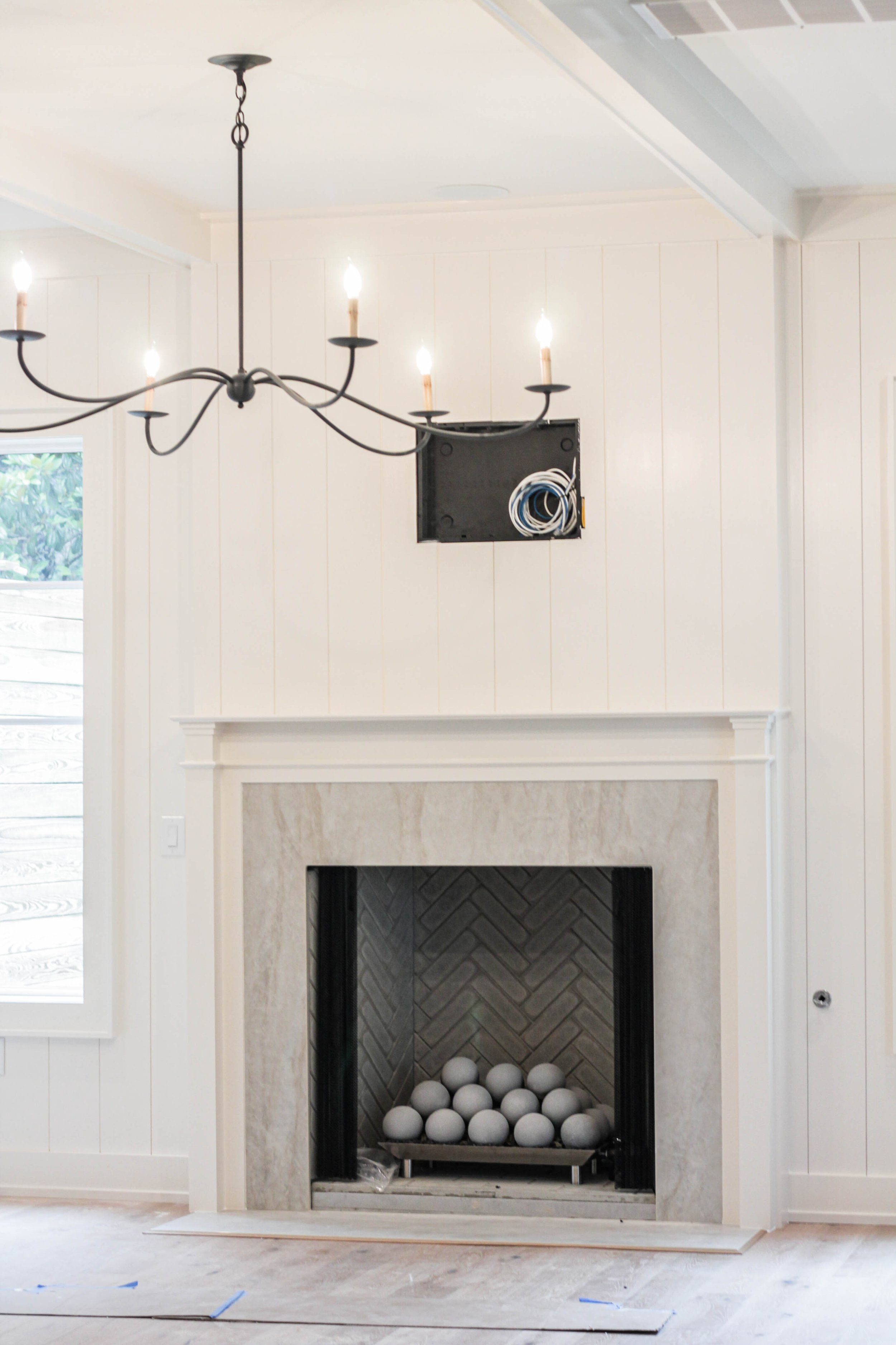 Living room with a fireplace under construction, black metal firebox with white balls inside, a white mantel, white paneled walls, a large window to the left, a chandelier hanging from the ceiling, and an open wall with visible wiring.