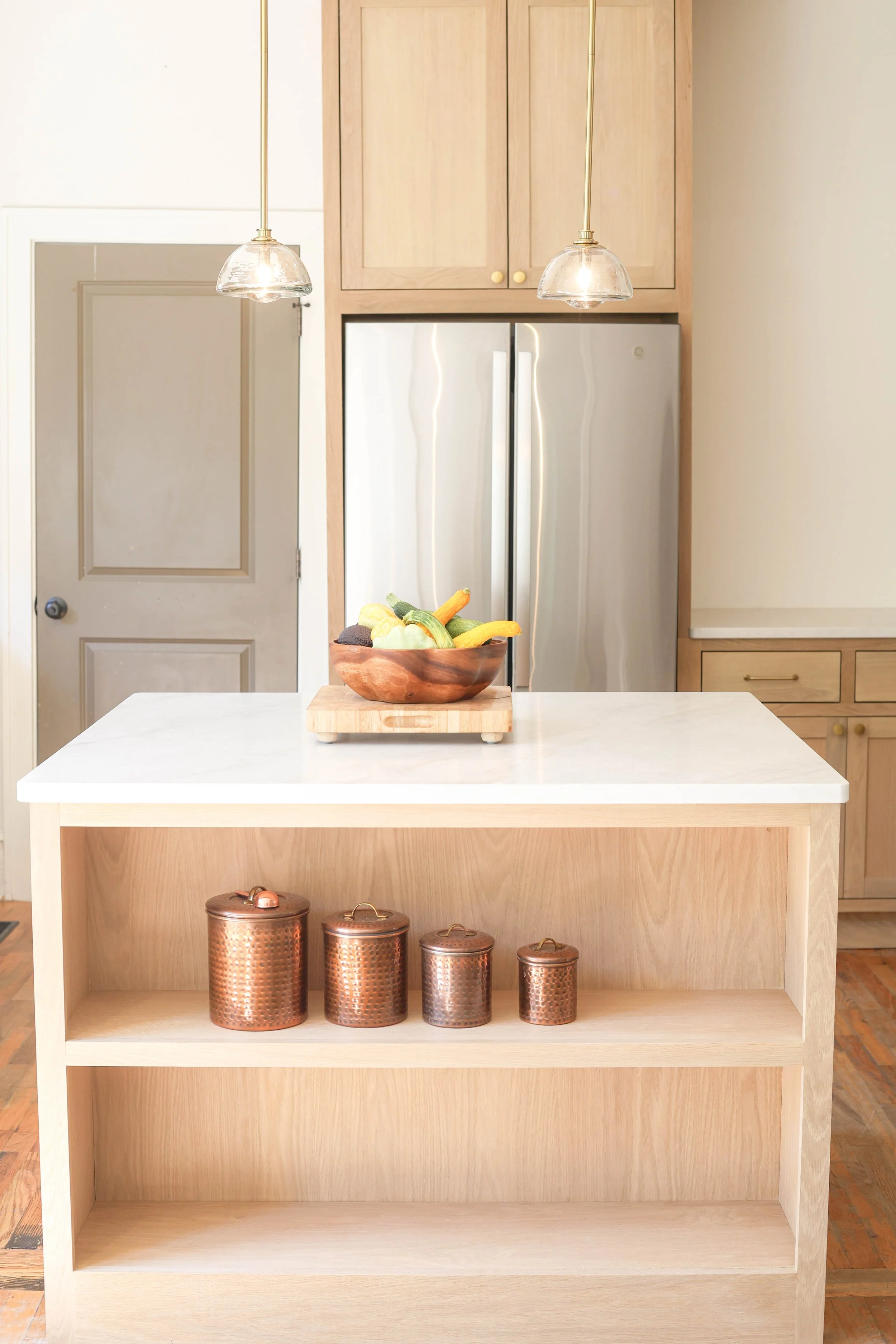 A kitchen island with a wooden bowl of assorted vegetables on top. Copper canisters on the lower shelf of the island. Light wood cabinetry and stainless steel refrigerator in the background. Two pendant lights hanging above the island.