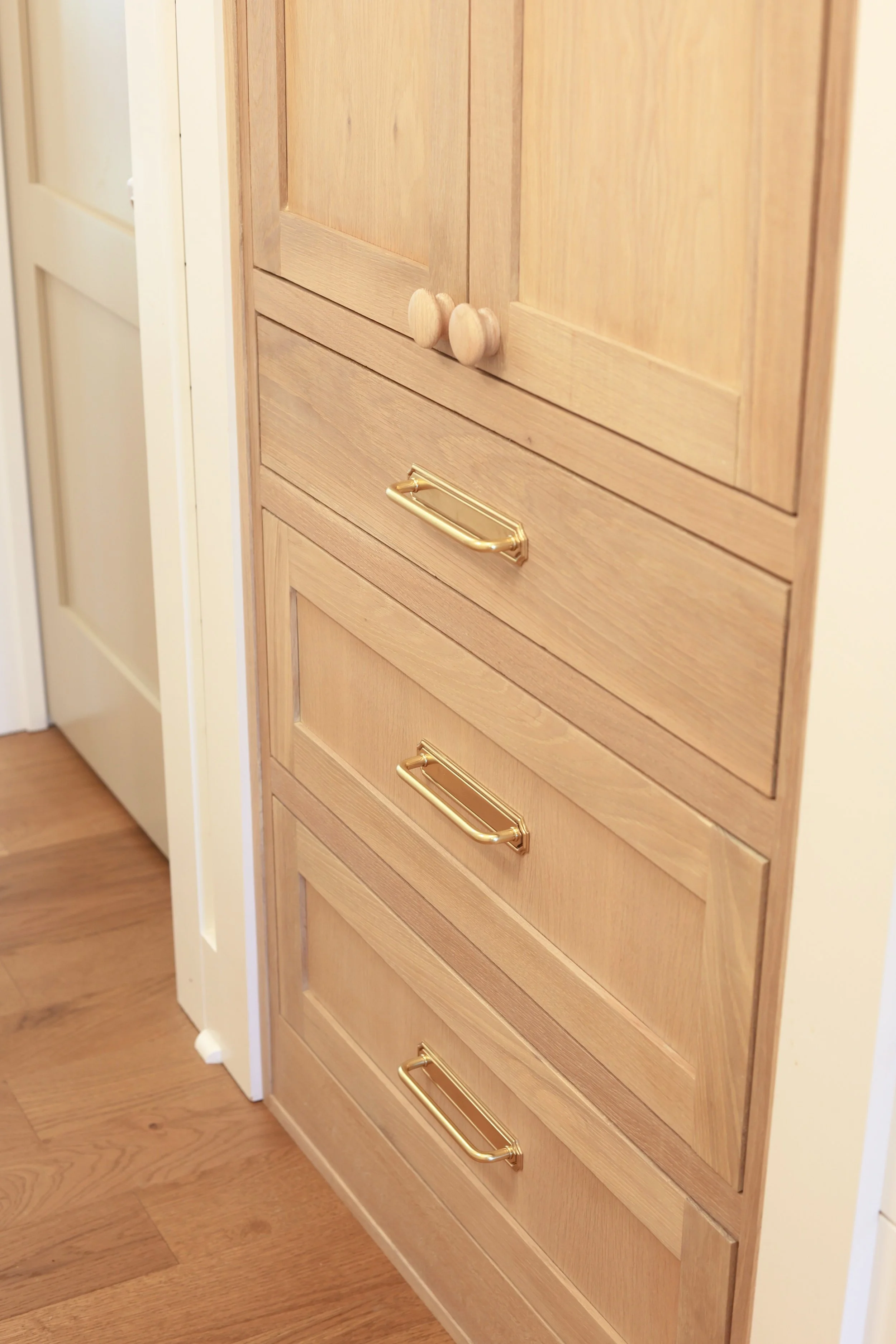 A wooden cabinet with three drawers, each equipped with gold-colored handles, positioned against a wooden floor and adjacent to a white door.